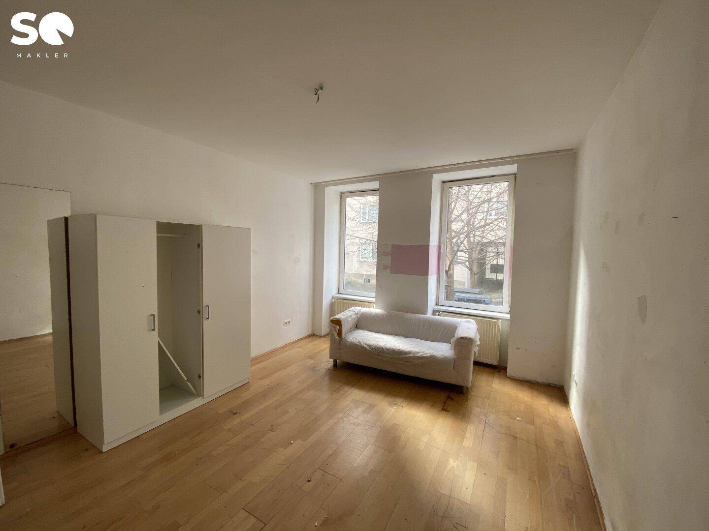 Heller Raum mit Holzboden, weißen Wänden, zwei Fenstern und einem kleinen Sofa.