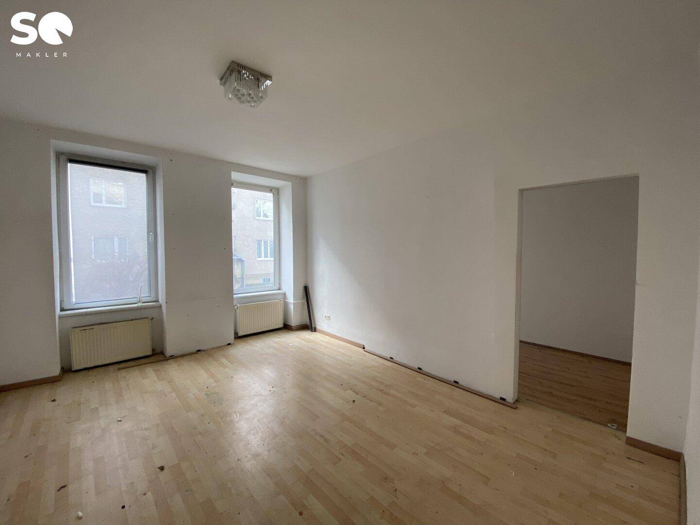 Leerer Raum mit Holzboden, weißen Wänden, zwei Fenstern und Zugang zu einem weiteren Zimmer.