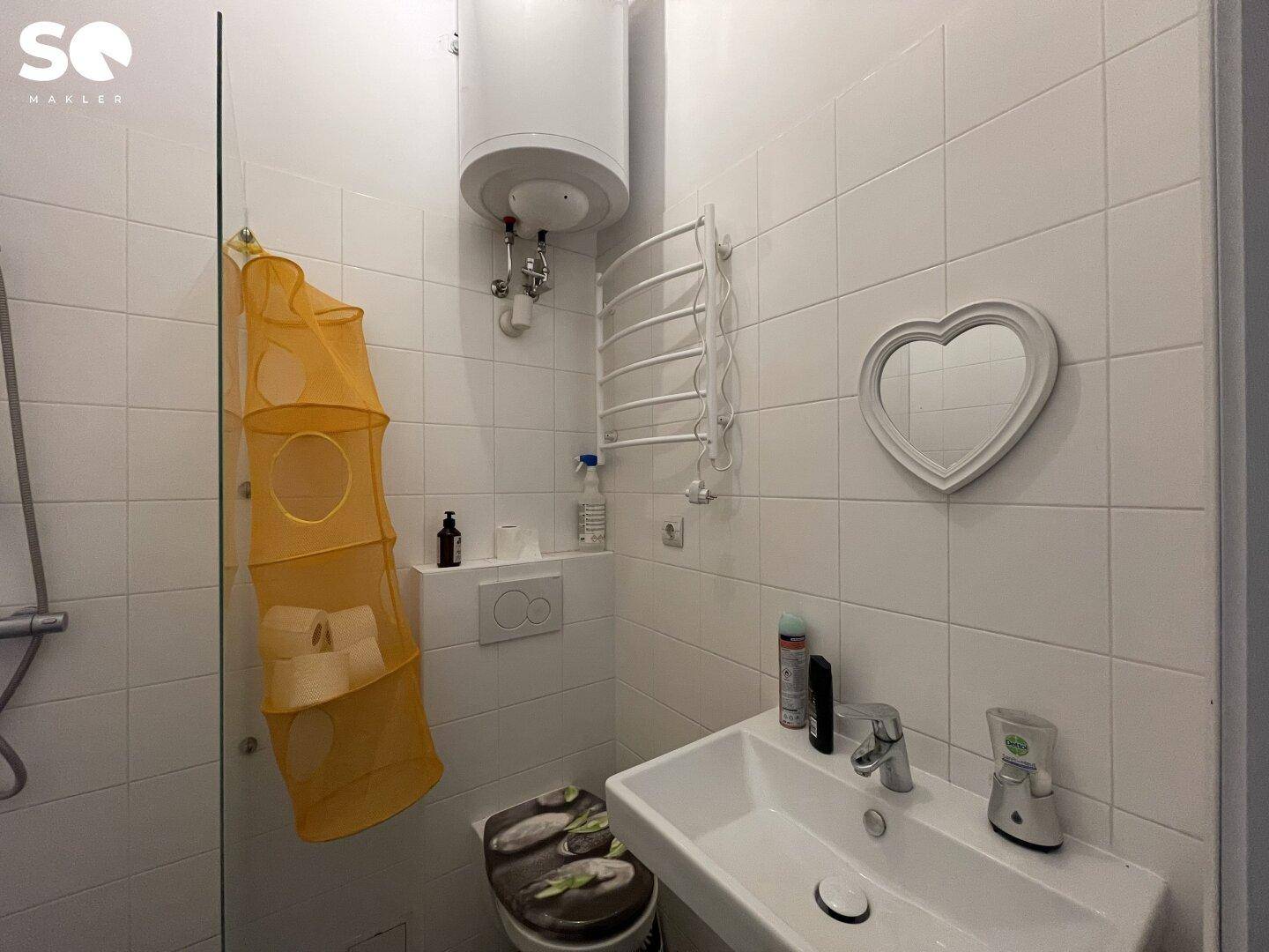 Helles Badezimmer mit weißer Fliesenwand, Waschbecken, WC und einem Warmwasserboiler.