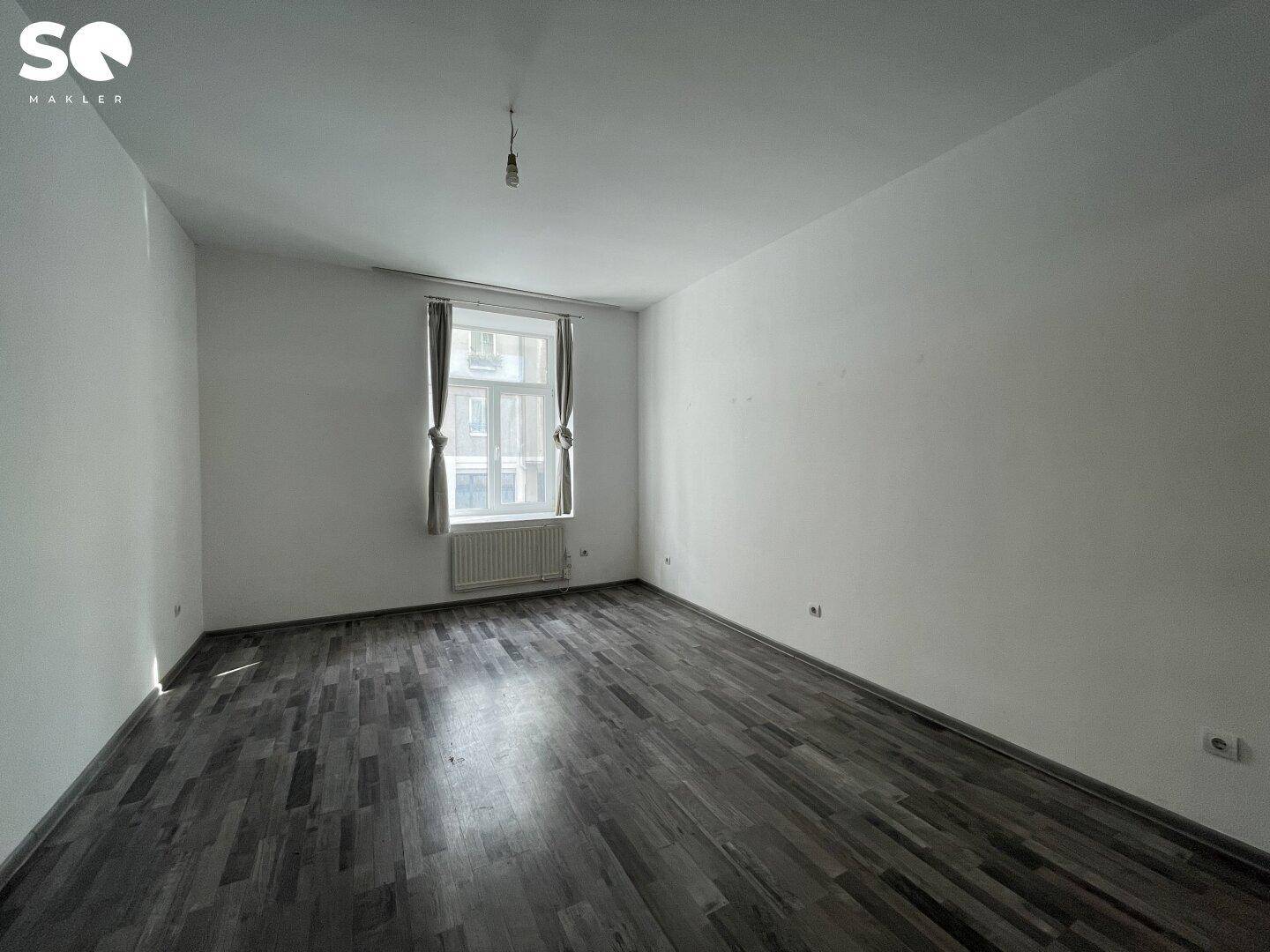 Geräumiger, heller Raum mit Laminatboden, einem Fenster und einem Heizkörper darunter.