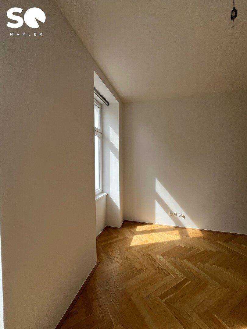 Lichtdurchfluteter Raum mit Fischgrätparkett und einem großen Fenster, das viel Tageslicht hereinlässt.