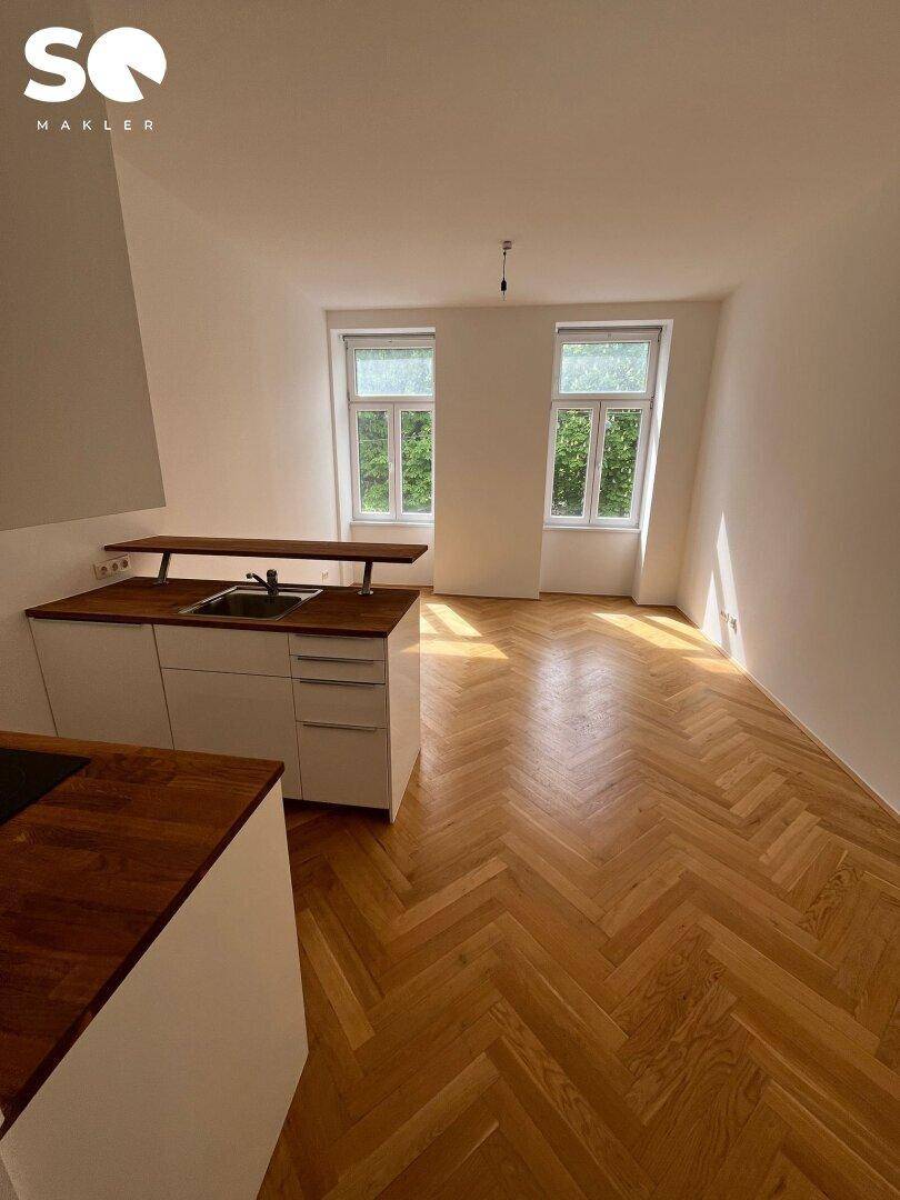Offener Wohnbereich mit integrierter Küche, Fischgrätparkett und Blick ins Grüne durch die Fenster.