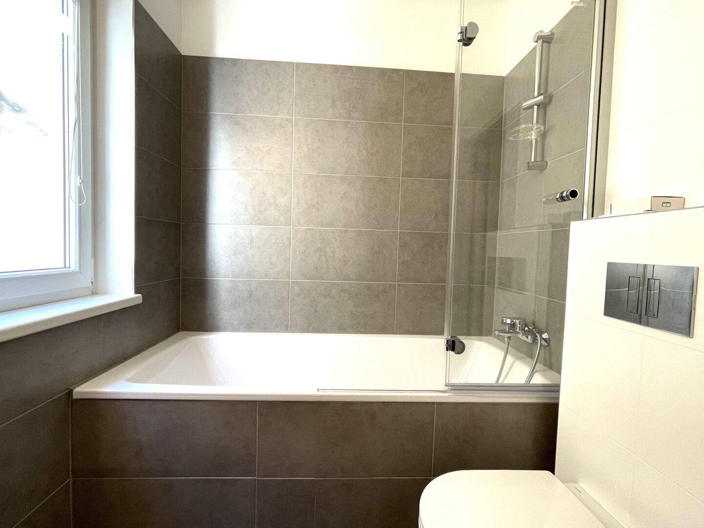 Modernes Badezimmer mit Badewanne, Duschabtrennung und grauen Fliesen.