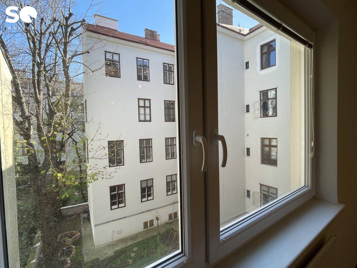 Blick aus dem Fenster auf einen ruhigen Innenhof mit Bäumen und angrenzenden Gebäuden.