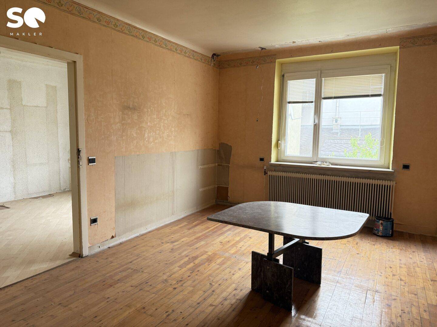 Renovierungsbedürftiger Raum mit Holzboden, Fenster und einem Heizkörper.