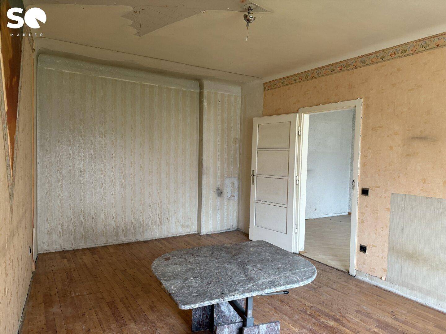 Renovierungsbedürftiger Raum mit altem Holzboden, abblätternder Tapete und einer Tür.