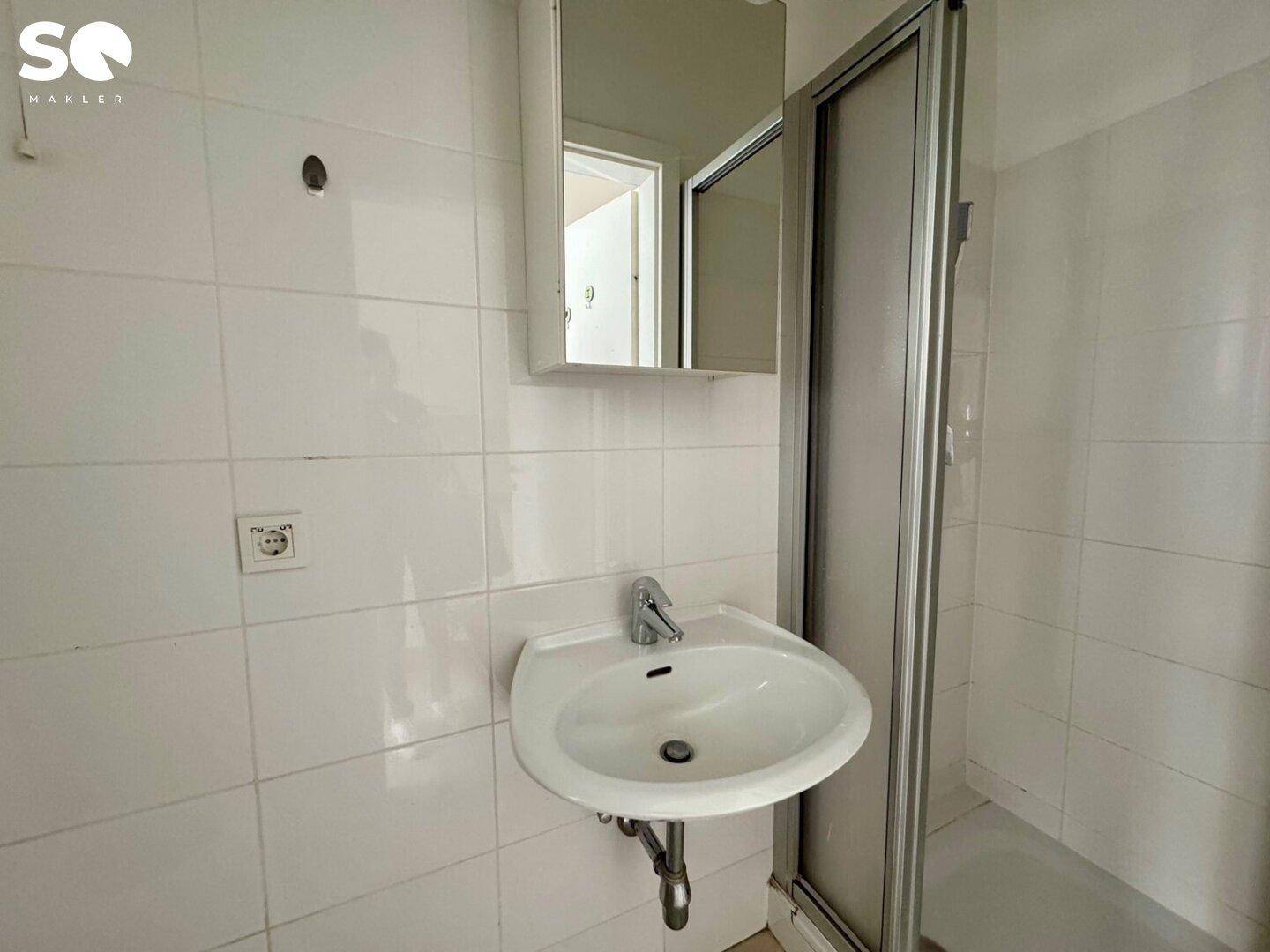 Badezimmer mit Waschbecken, Spiegelschrank und Duschkabine, komplett weiß gefliest.