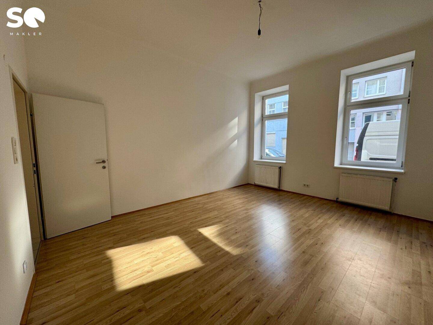 Lichtdurchflutetes Zimmer mit Holzfußboden und zwei Fenstern, ideal für Wohnzwecke.