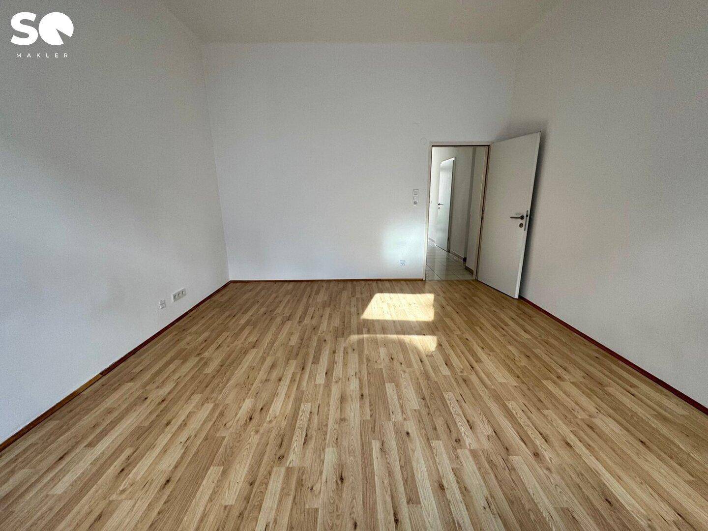 Geräumiges, leeres Zimmer mit Holzfußboden und Zugang zu einem hellen Flur.
