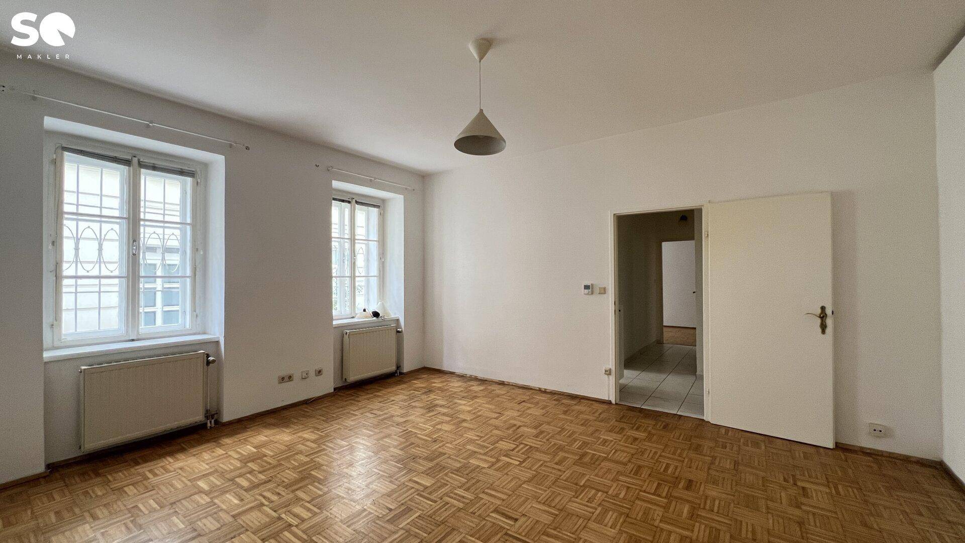 Geräumiges Wohnzimmer mit Parkettboden und Zugang zu einem weiteren Raum.