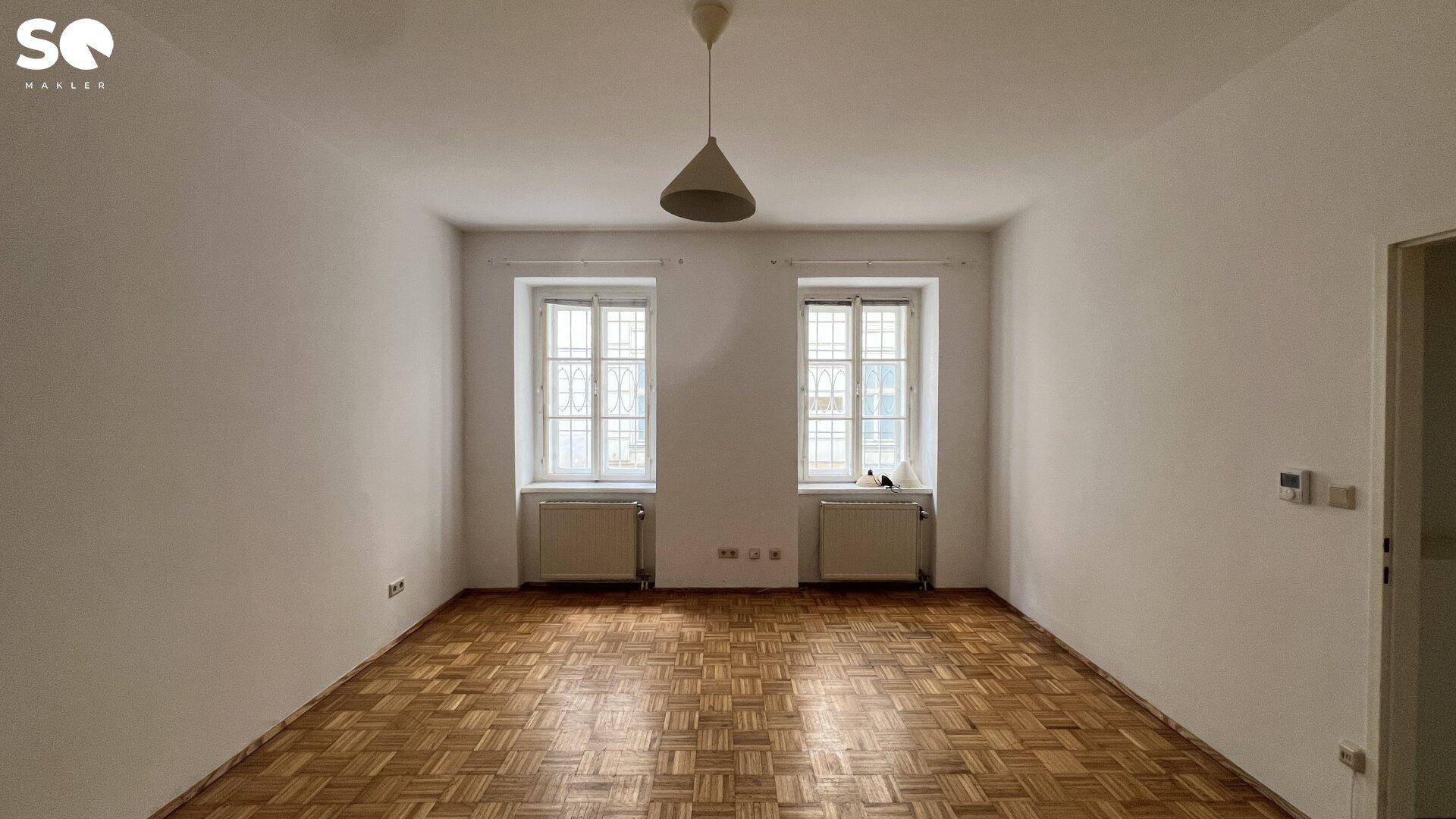Lichtdurchflutetes Zimmer mit klassischem Parkettboden und zwei großen Fenstern.