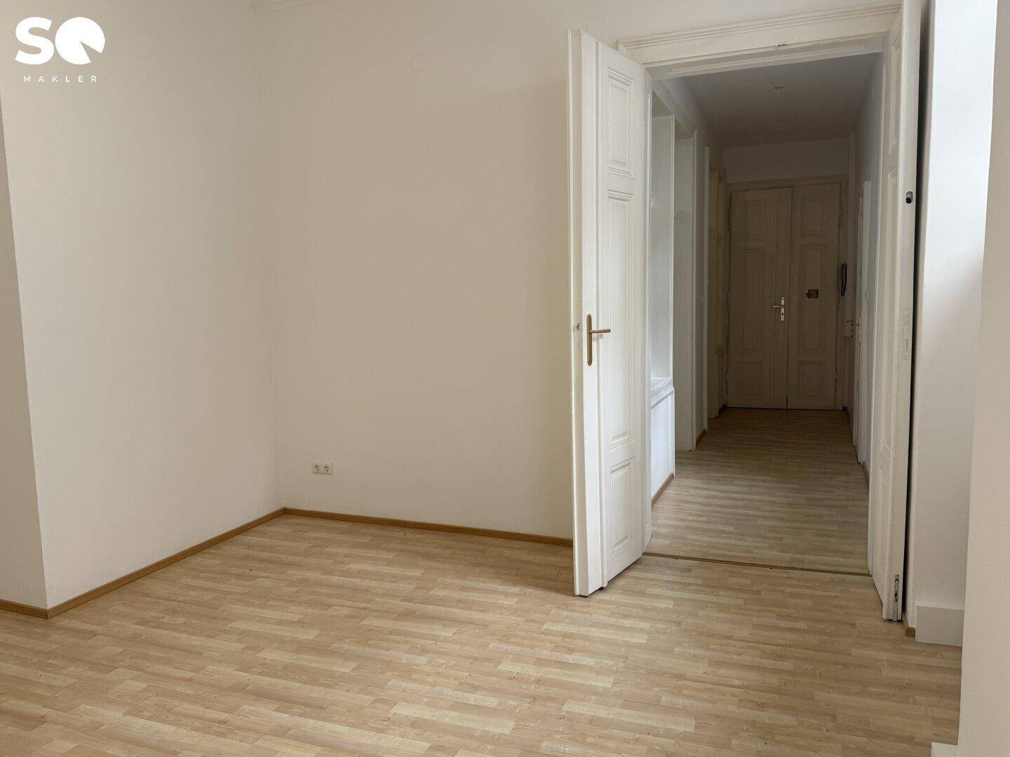 Geräumiges, leeres Zimmer mit Holzboden und Zugang zu einem langen Flur.