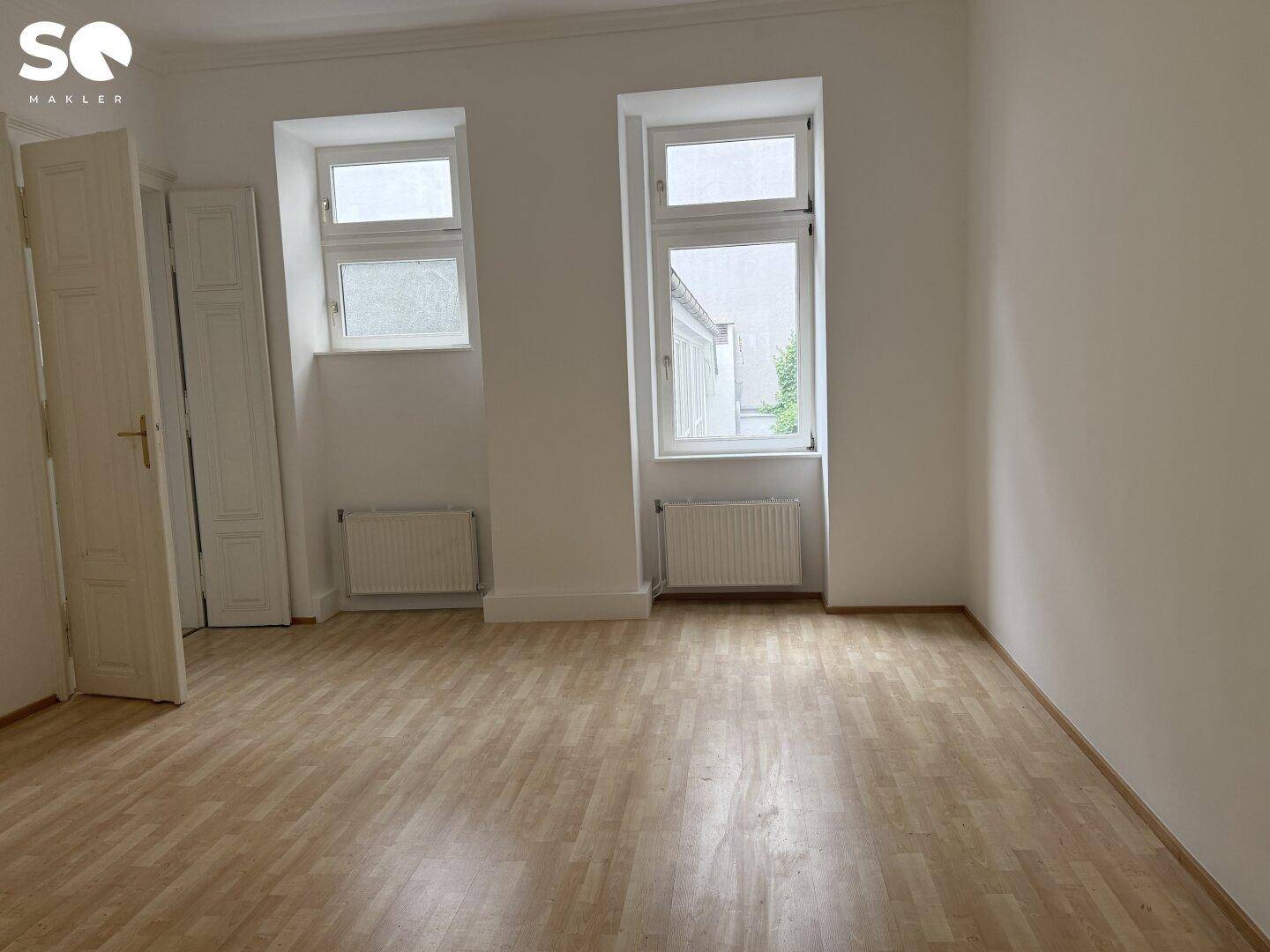Helles, leeres Zimmer mit Holzboden und zwei Fenstern zum Innenhof.