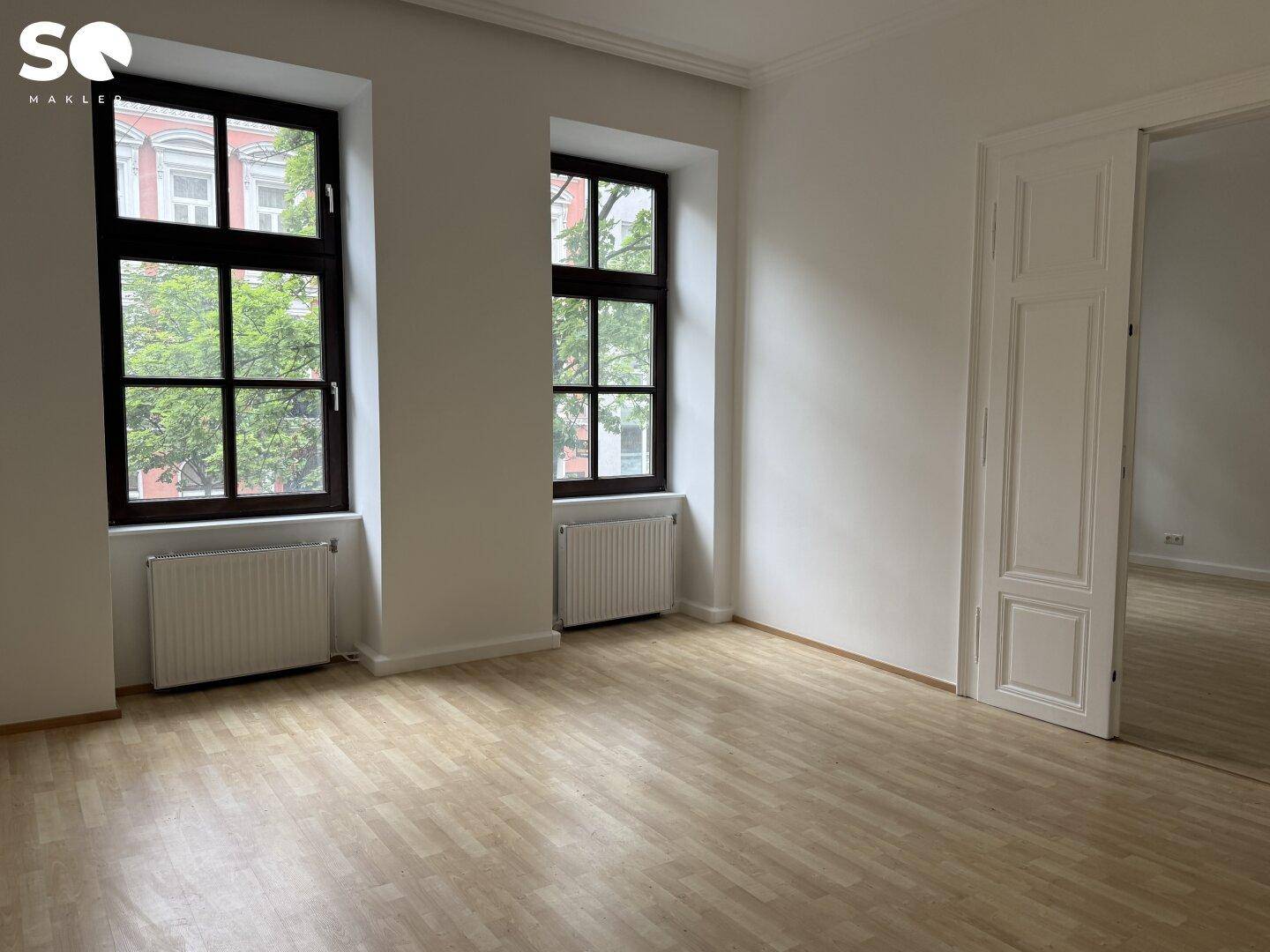 Helles, leeres Zimmer mit Holzboden, zwei Fenstern und einem Durchgang.