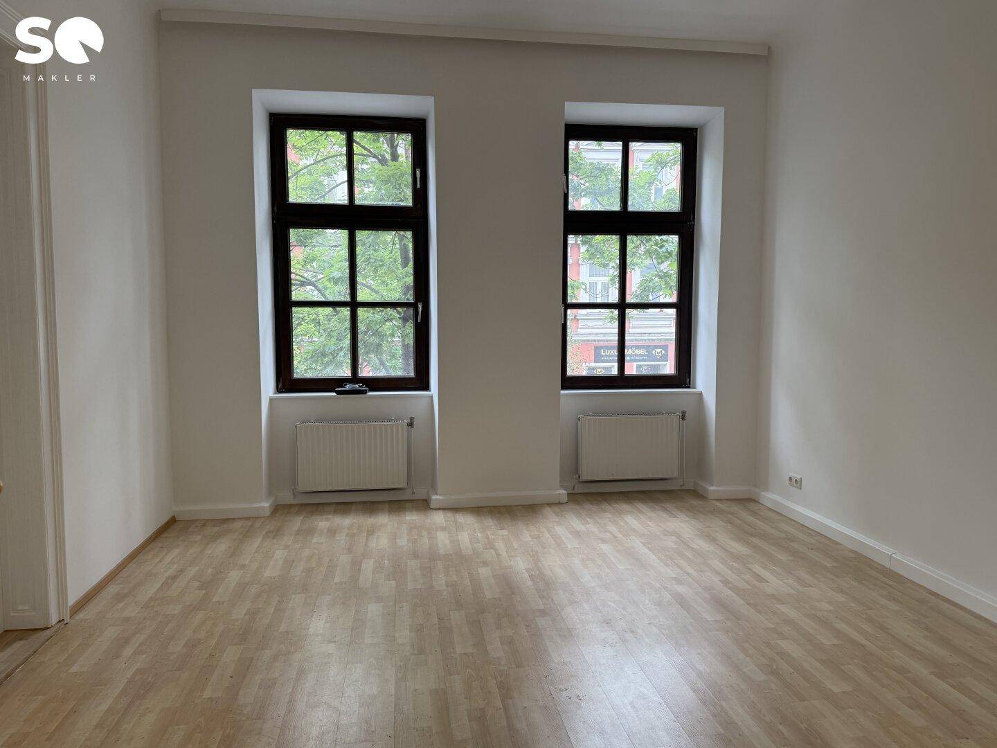 Helles, leeres Zimmer mit Holzboden und zwei Fenstern zur Straße.