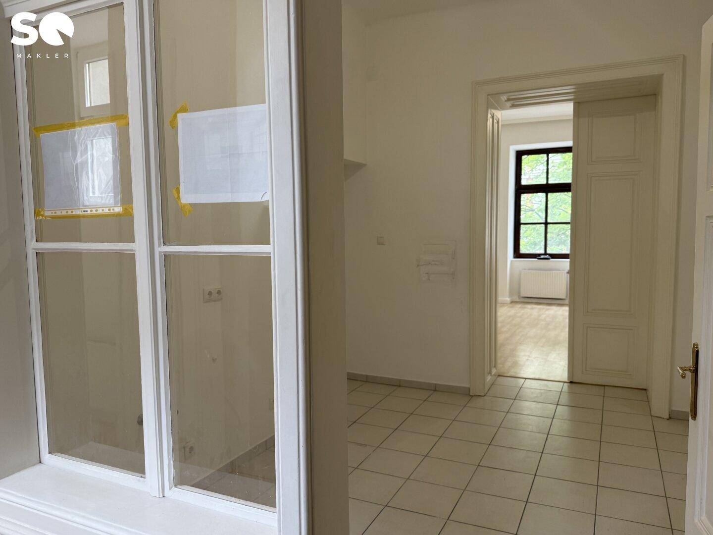 Raum mit Fliesenboden und Durchgang zu einem hellen Zimmer mit Holzboden.