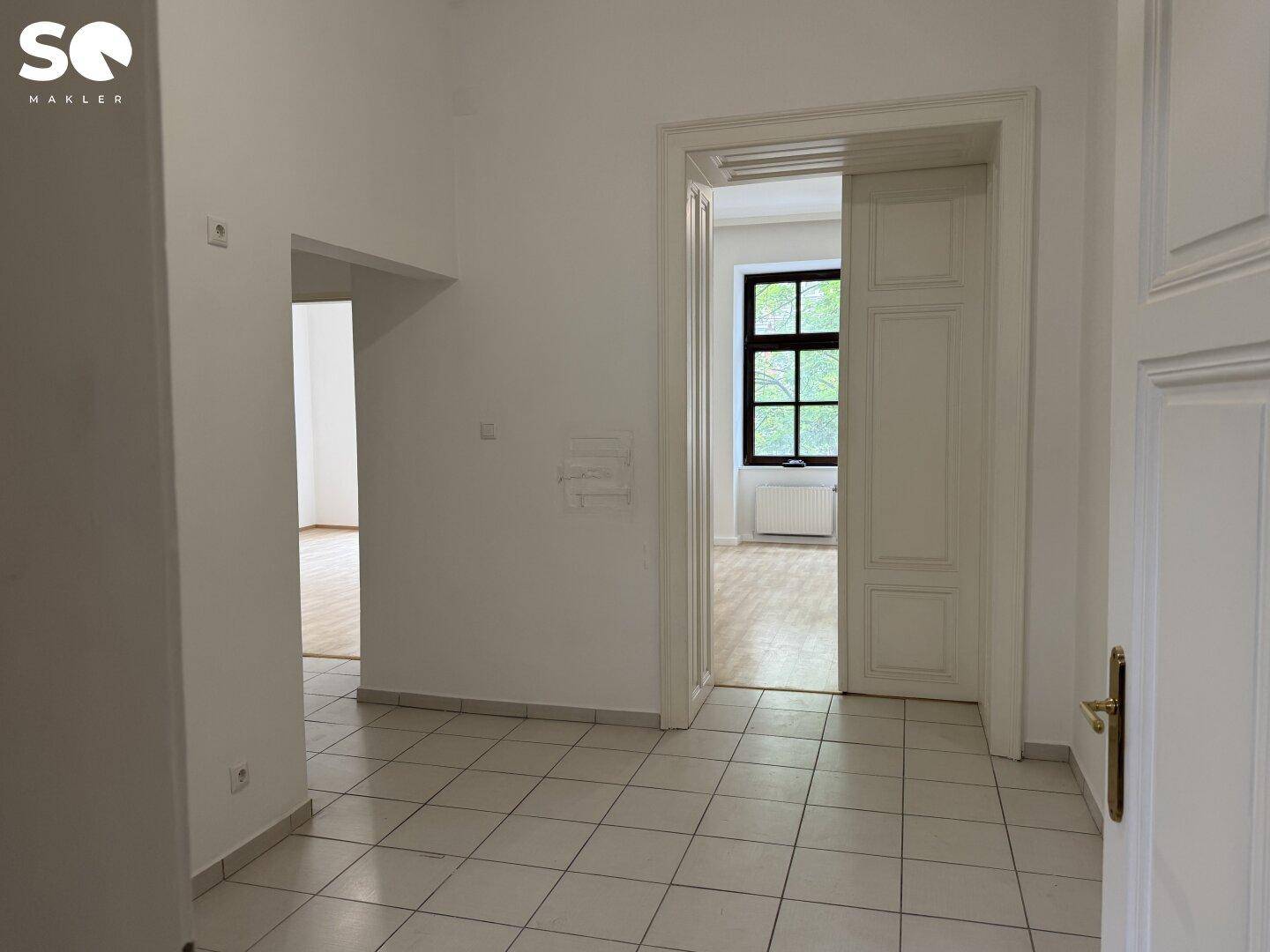 Heller Eingangsbereich mit Fliesenboden und Durchgang zu einem Zimmer mit Holzboden.