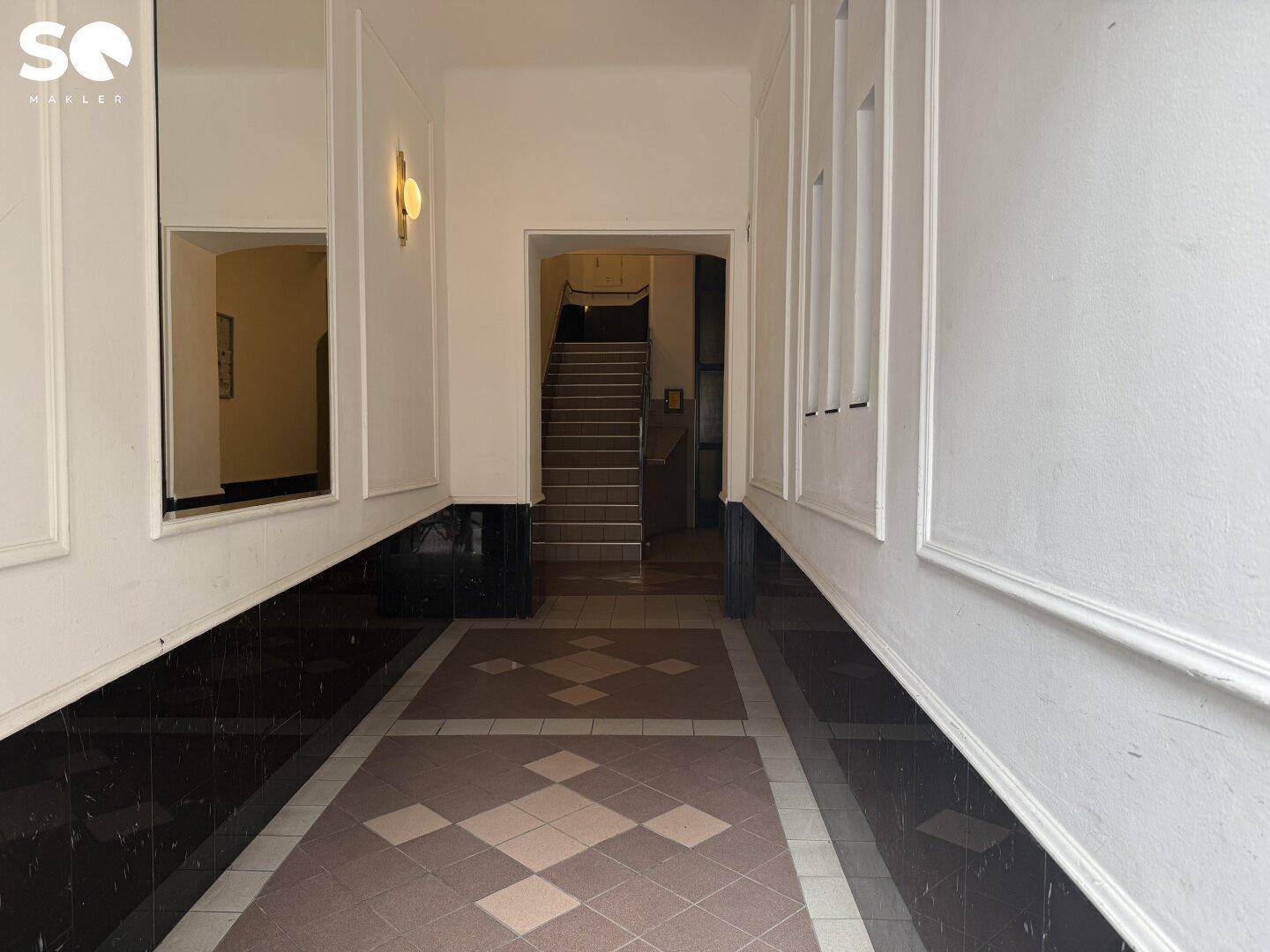 Langer Flur mit gemusterten Fliesen und dekorativen Wandverkleidungen, der zum Treppenhaus führt.