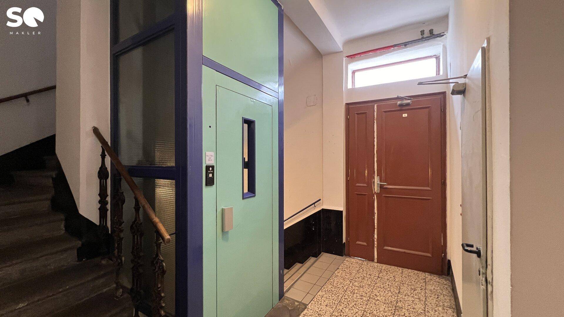 Eingangsbereich mit einem modernen Aufzug und einer Treppe, die den Zugang zu den oberen Etagen ermöglicht.