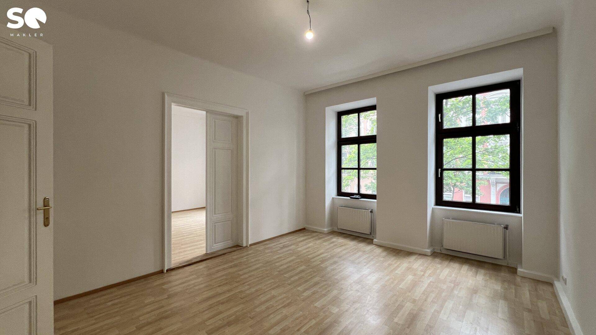Geräumiges, leeres Zimmer mit Holzboden, hohen Decken und zwei großen Fenstern.