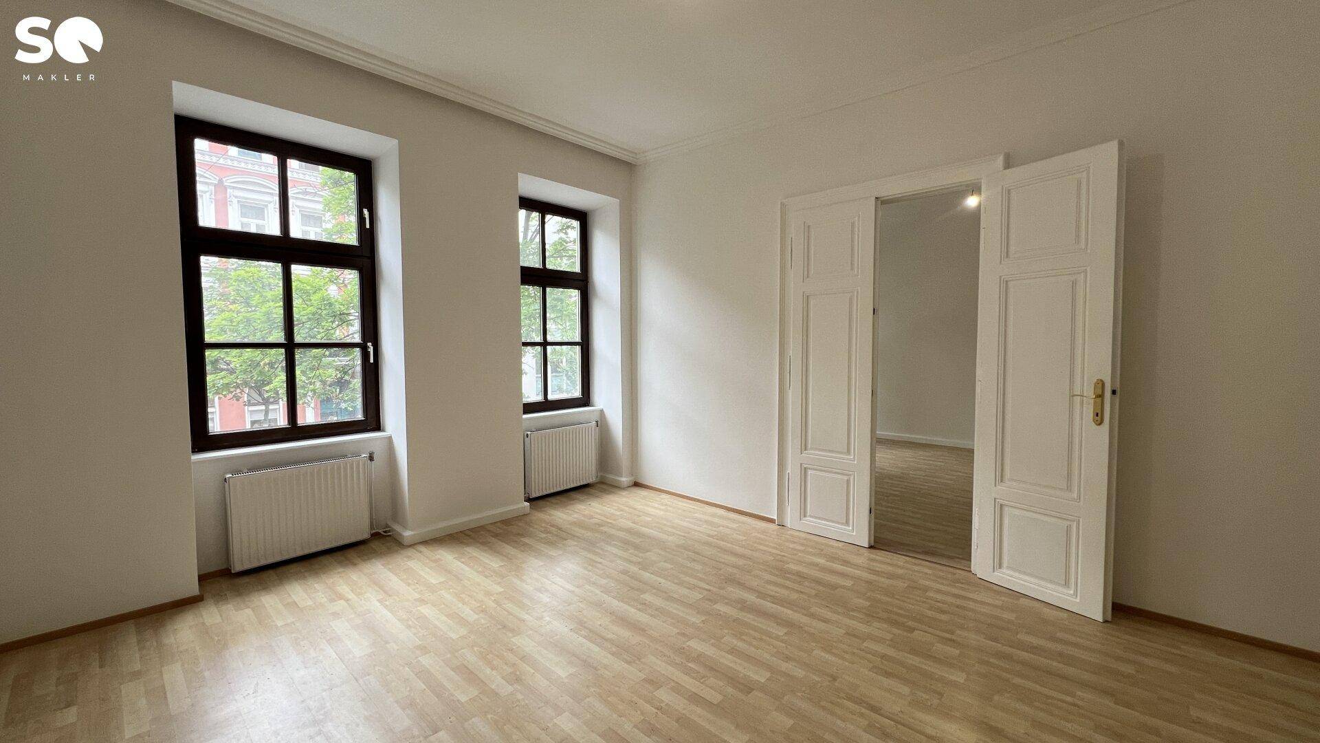 Helles, leeres Zimmer mit Holzboden, zwei Fenstern und einem Durchgang zu einem weiteren Raum.