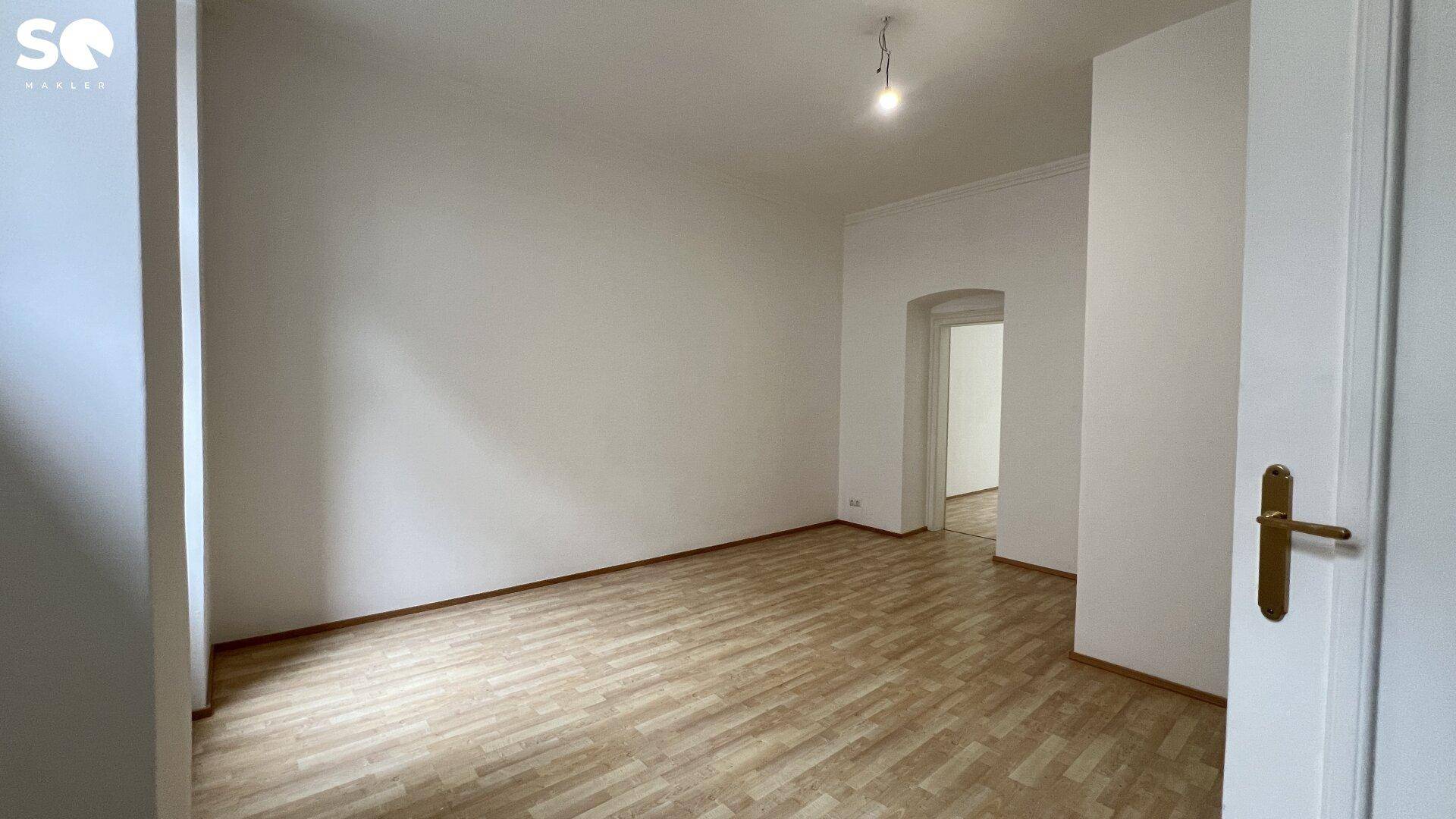 Geräumiges, leeres Zimmer mit Holzboden und einem Durchgang zu einem weiteren Raum.