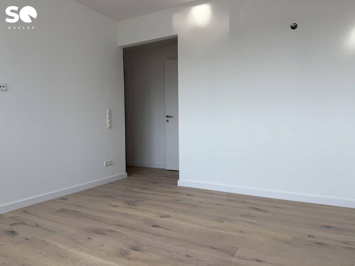 Leerer Raum mit hellem Holzboden und weißen Wänden, der zu einem weiteren Zimmer führt.