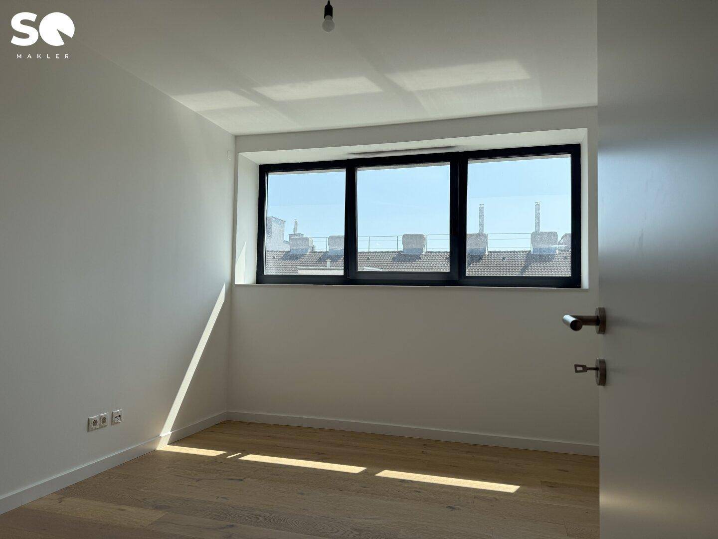 Geräumiges Schlafzimmer mit hellem Holzboden und einem Fenster mit Blick auf die Dächer der Stadt.