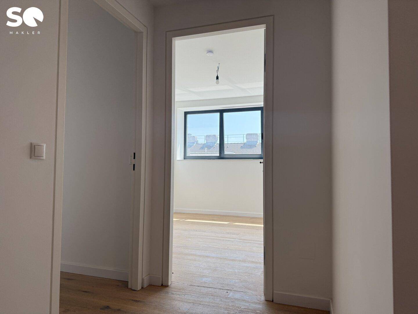 Blick aus einem Flur in ein helles Zimmer mit Holzboden und einem Fenster mit Fernsicht.