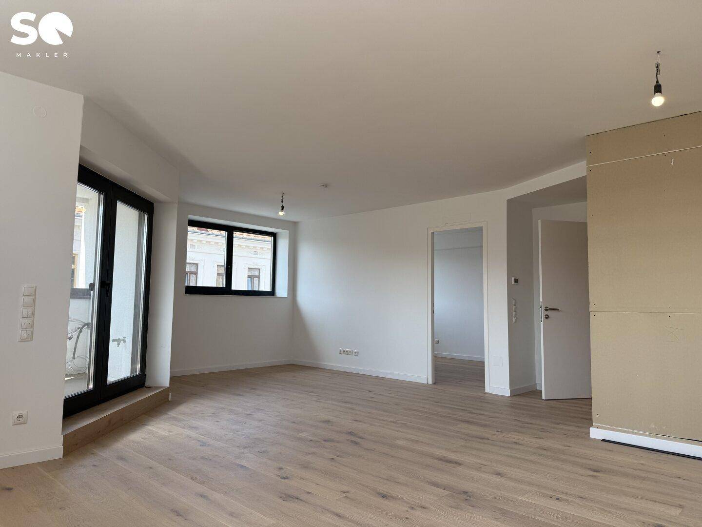 Geräumiges Wohnzimmer mit hellem Holzboden, weißen Wänden und Zugang zu einem Balkon.
