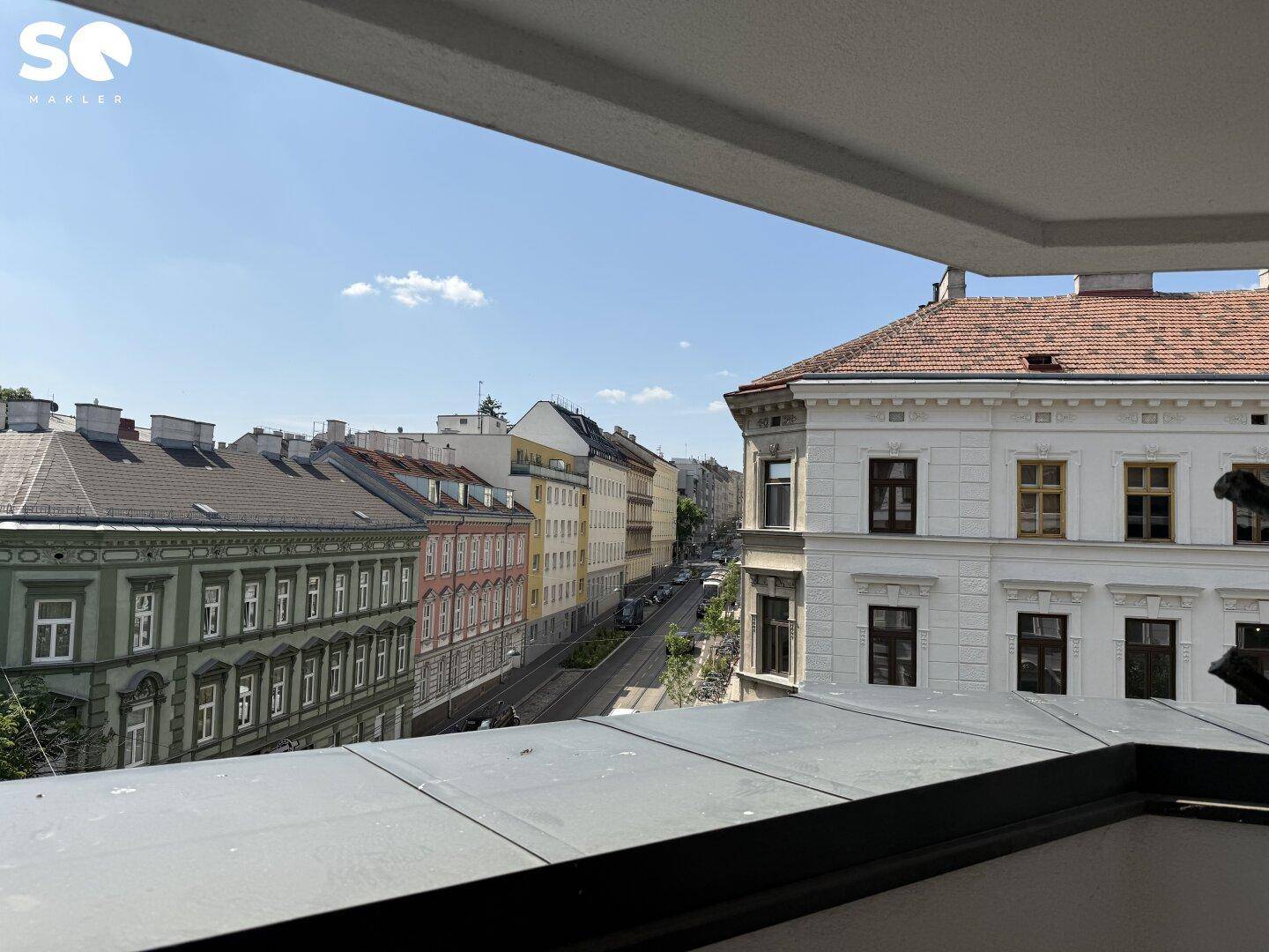 Blick von einem Balkon auf eine belebte Stadtstraße mit historischen Gebäuden und blauem Himmel.