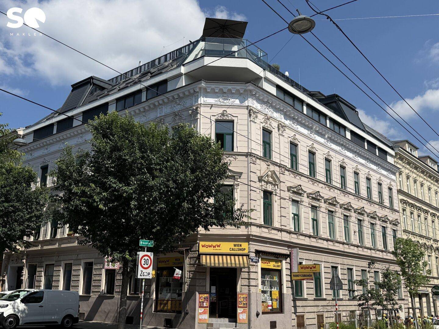 Eckgebäude mit historischer Fassade und modernem Dachausbau, umgeben von Bäumen und städtischer Umgebung.