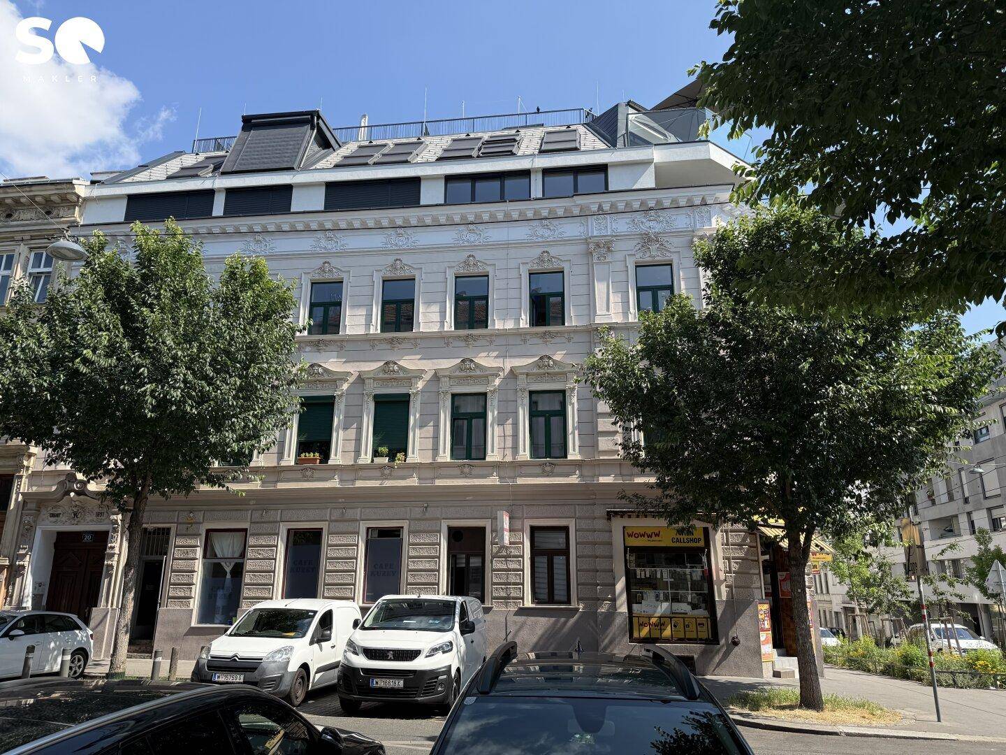 Ansicht der historischen Fassade des Gebäudes mit modernem Dachausbau, umgeben von parkenden Autos und Bäumen.
