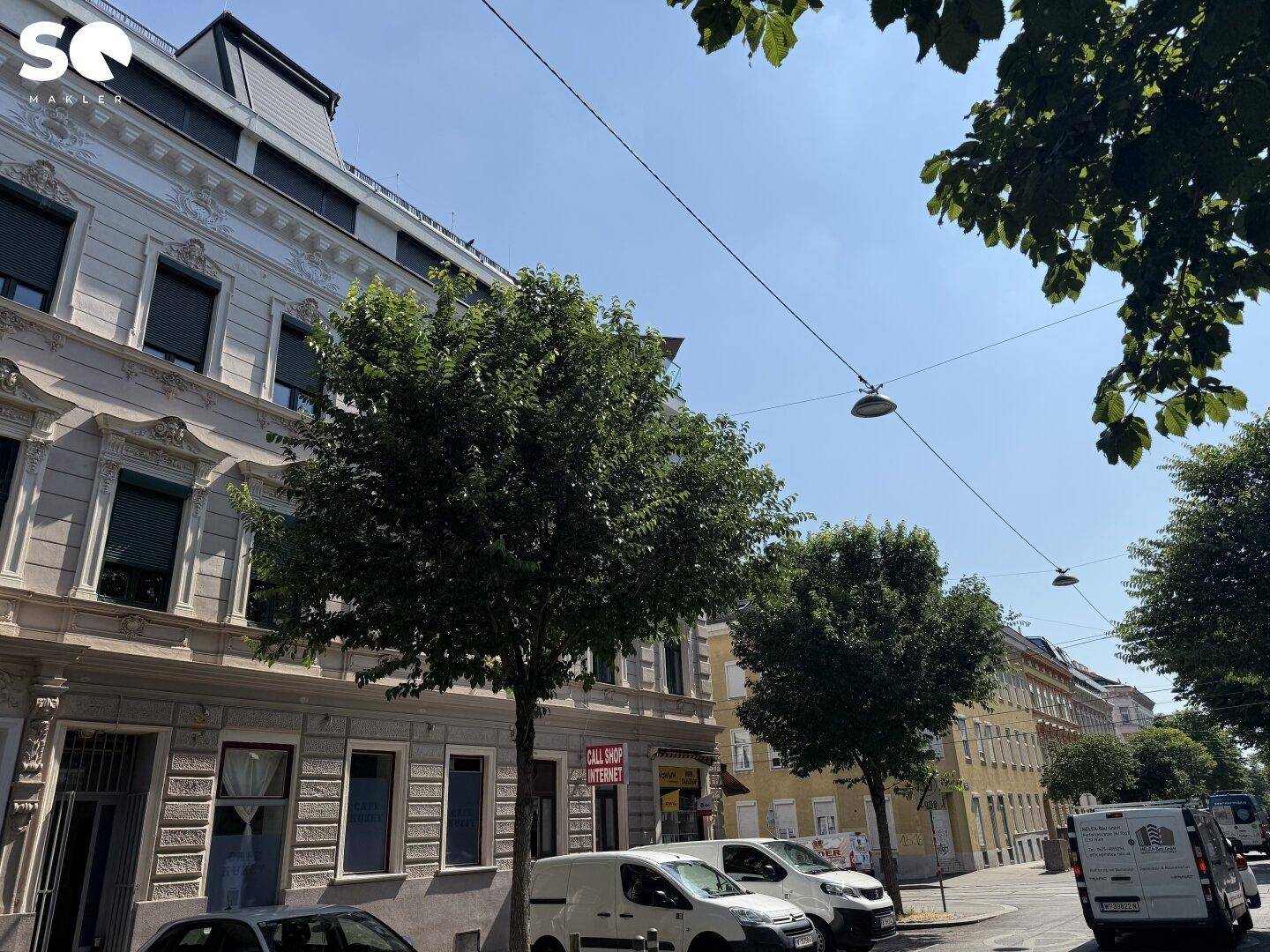 Straßenszene mit historischem Gebäude, Bäumen und parkenden Fahrzeugen unter blauem Himmel.