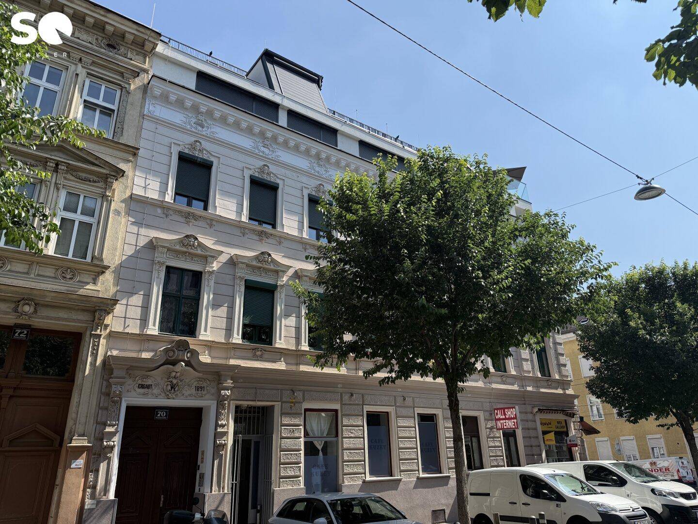 Historisches Gebäude mit modernem Dachausbau und Bäumen im Vordergrund, an einer belebten Straße gelegen.