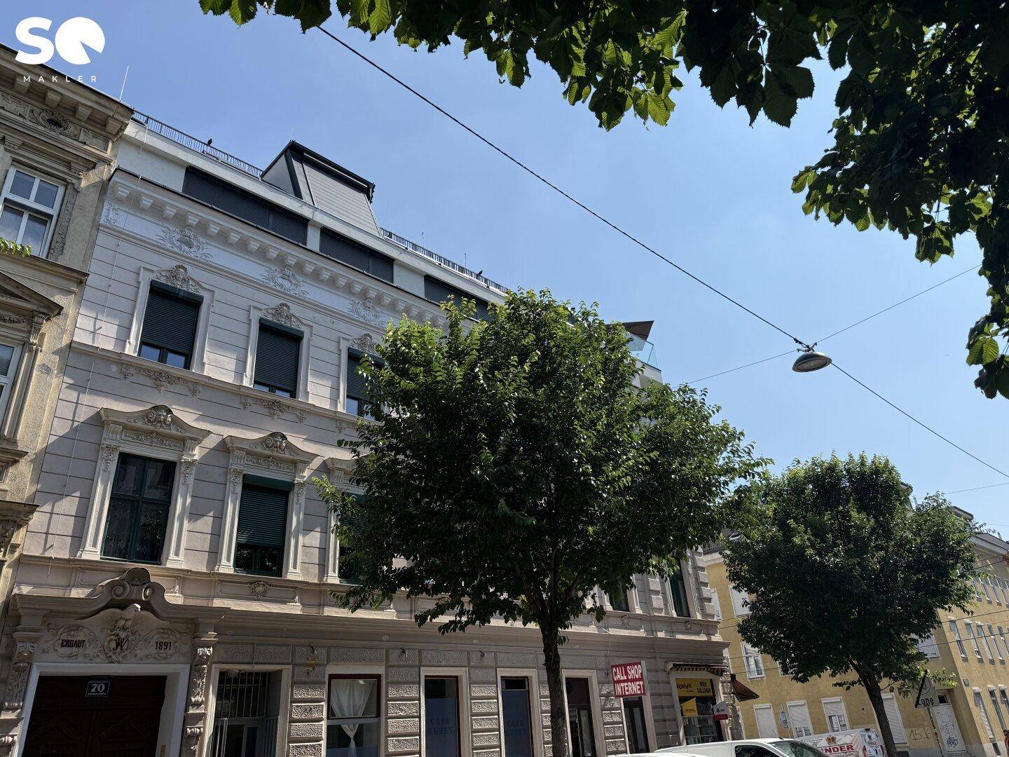 Straßenszene mit historischem Gebäude, Bäumen und klarem Himmel, das moderne Dachgeschoss ist sichtbar.