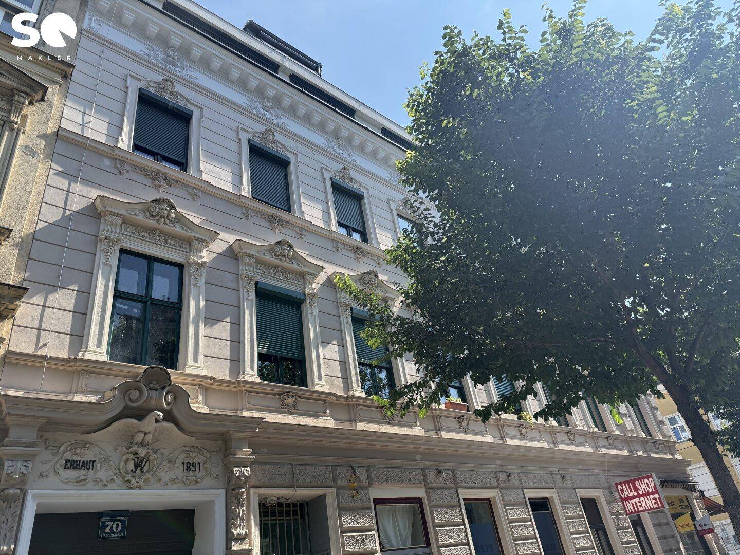 Detailansicht der historischen Fassade mit aufwendigen Verzierungen und dem Baujahr 1891, teilweise von Baum verdeckt.