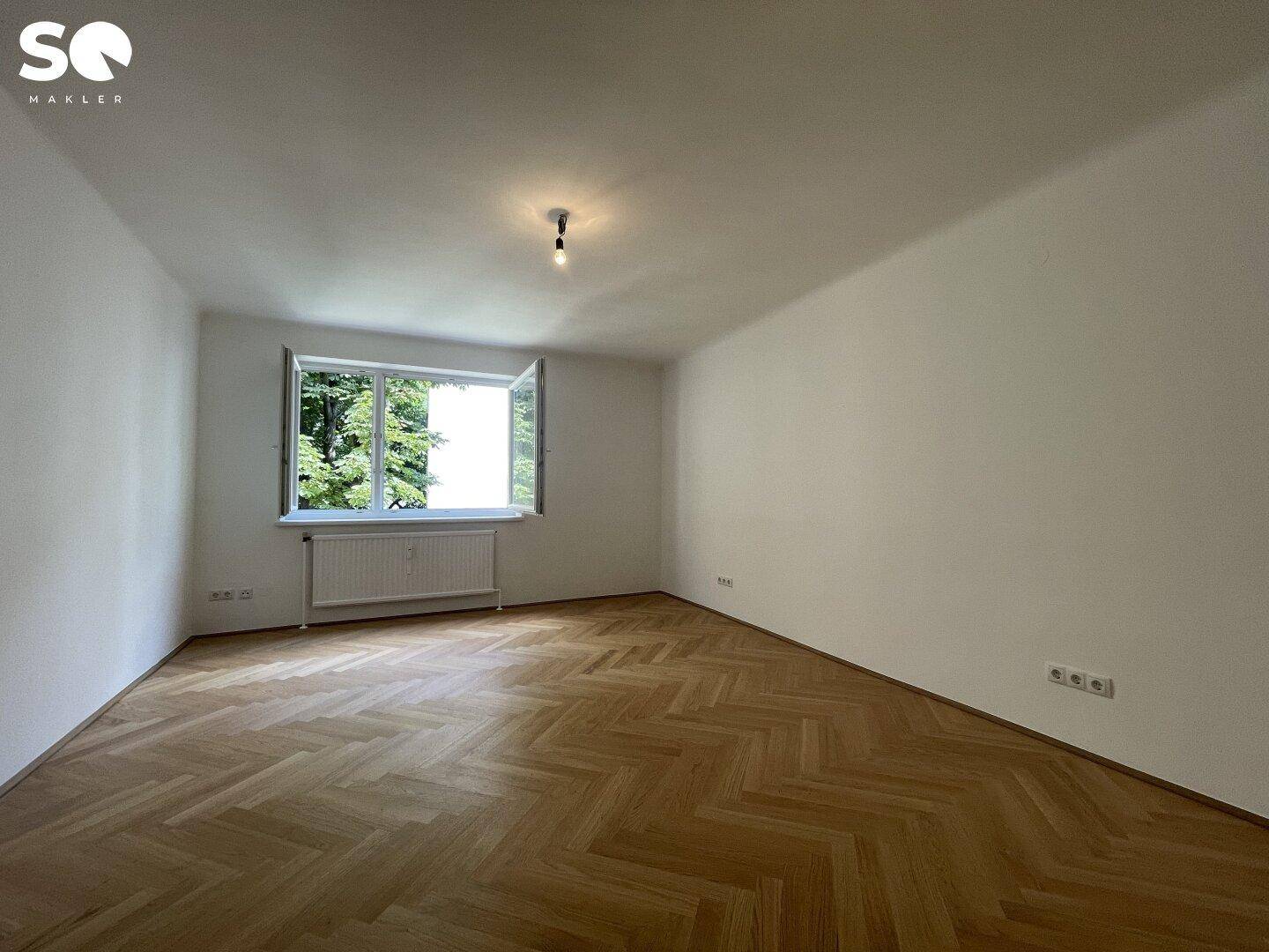 Geräumiges Zimmer mit Fischgrätparkett und Blick ins Grüne durch das geöffnete Fenster.