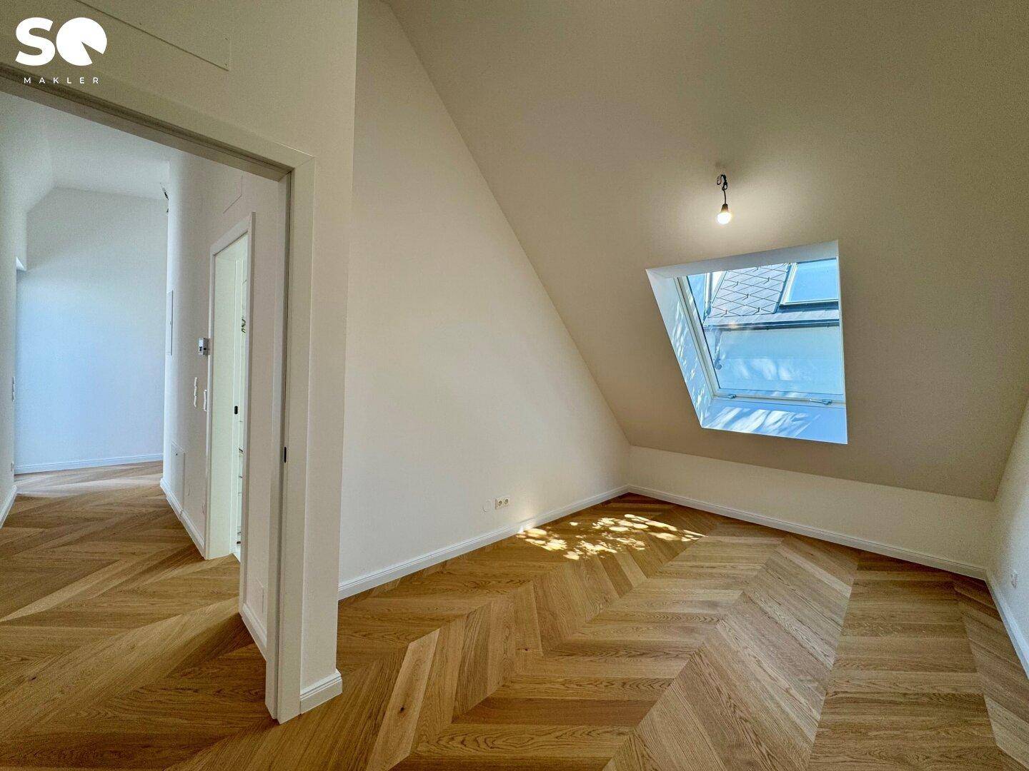 Helles Zimmer im Dachgeschoss mit Fischgrätparkett, weißen Wänden und einem großen Dachfenster.
