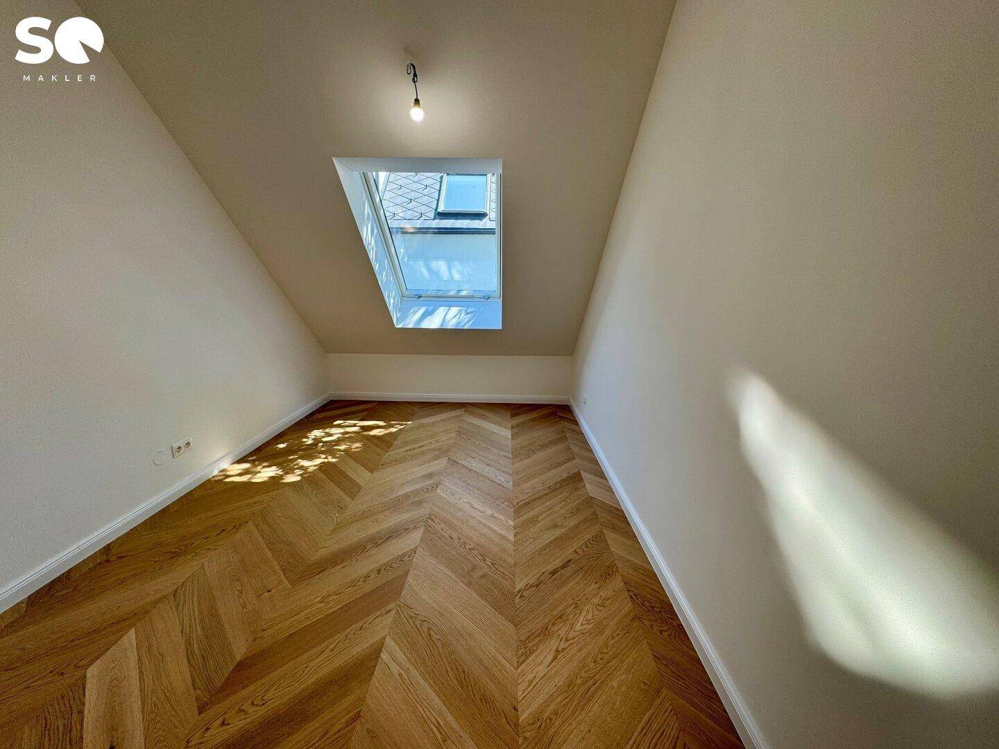 Helles Dachzimmer mit schrägem Dach und Fischgrätparkettboden, beleuchtet durch ein Dachfenster.