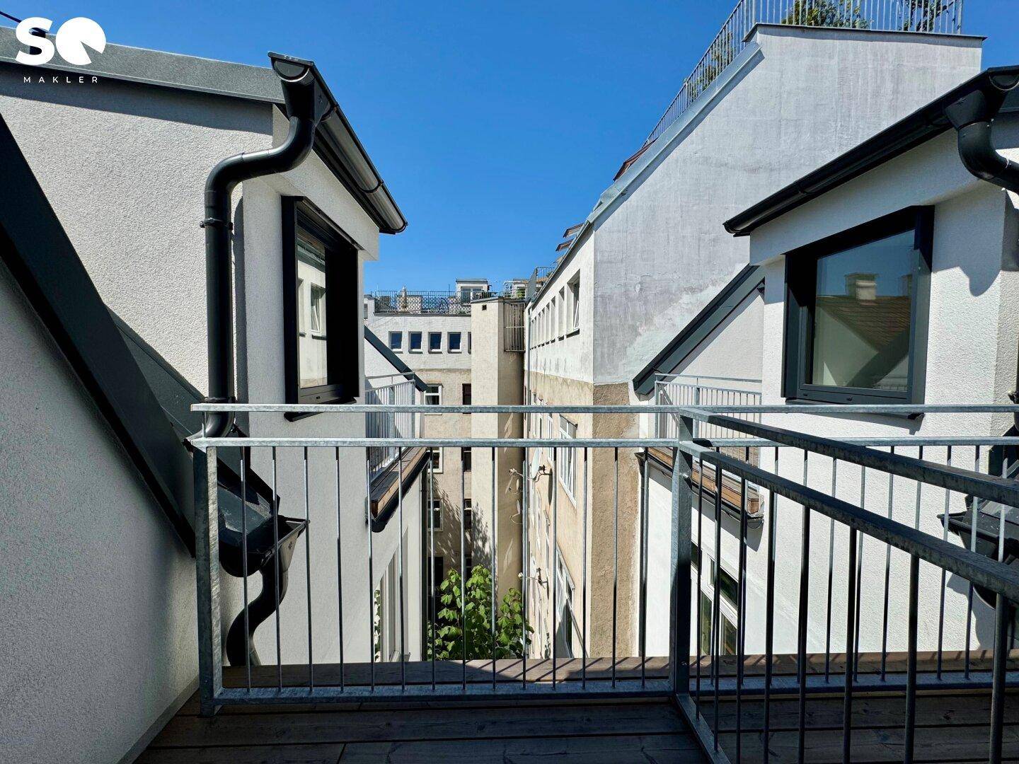 Balkon mit Holzboden und Metallgeländer, der einen Blick auf die benachbarten Gebäude und den Himmel freigibt.