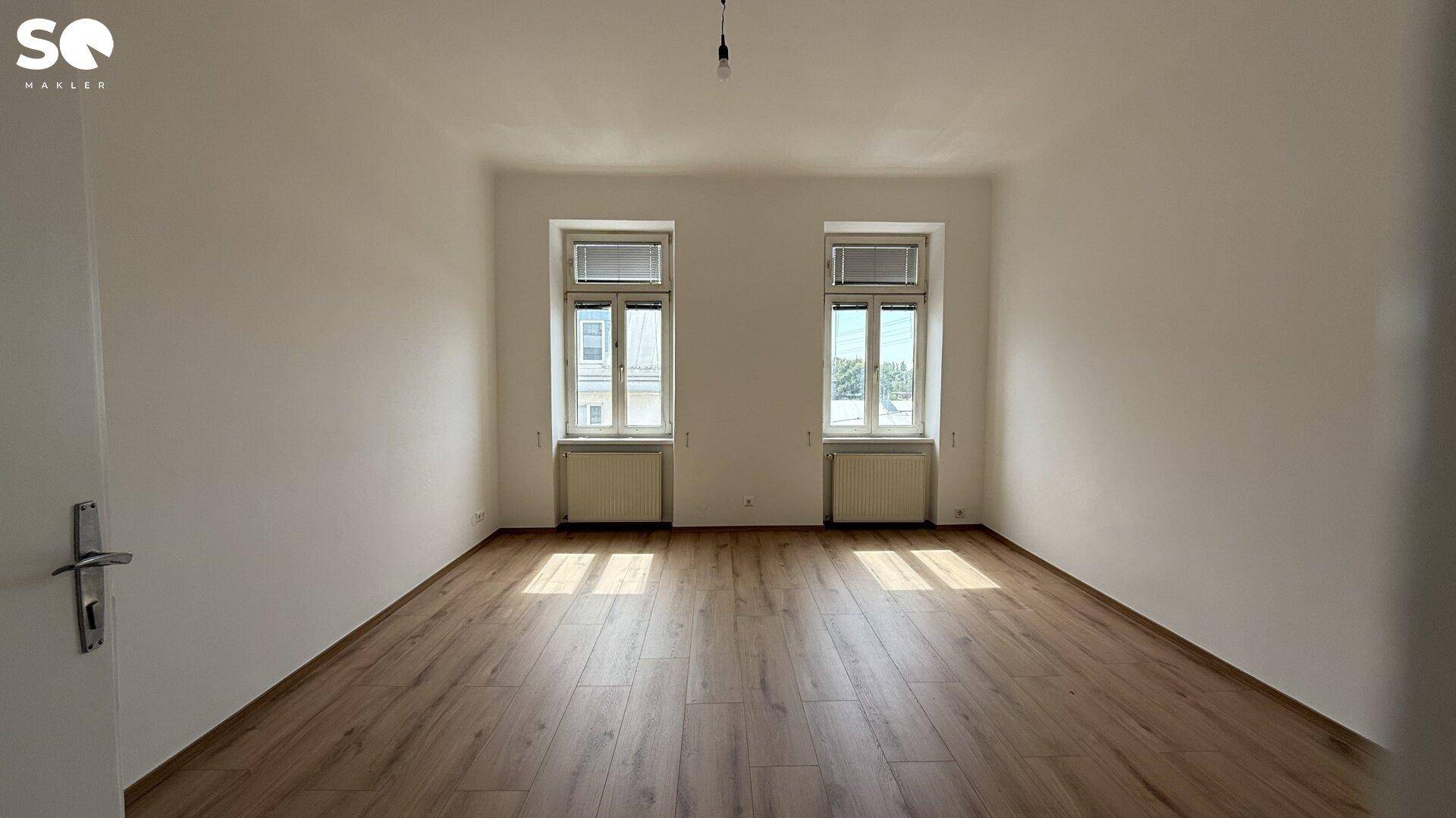 Freundliches, unmöbliertes Zimmer mit Holzboden und zwei großen Fenstern, viel Tageslicht.