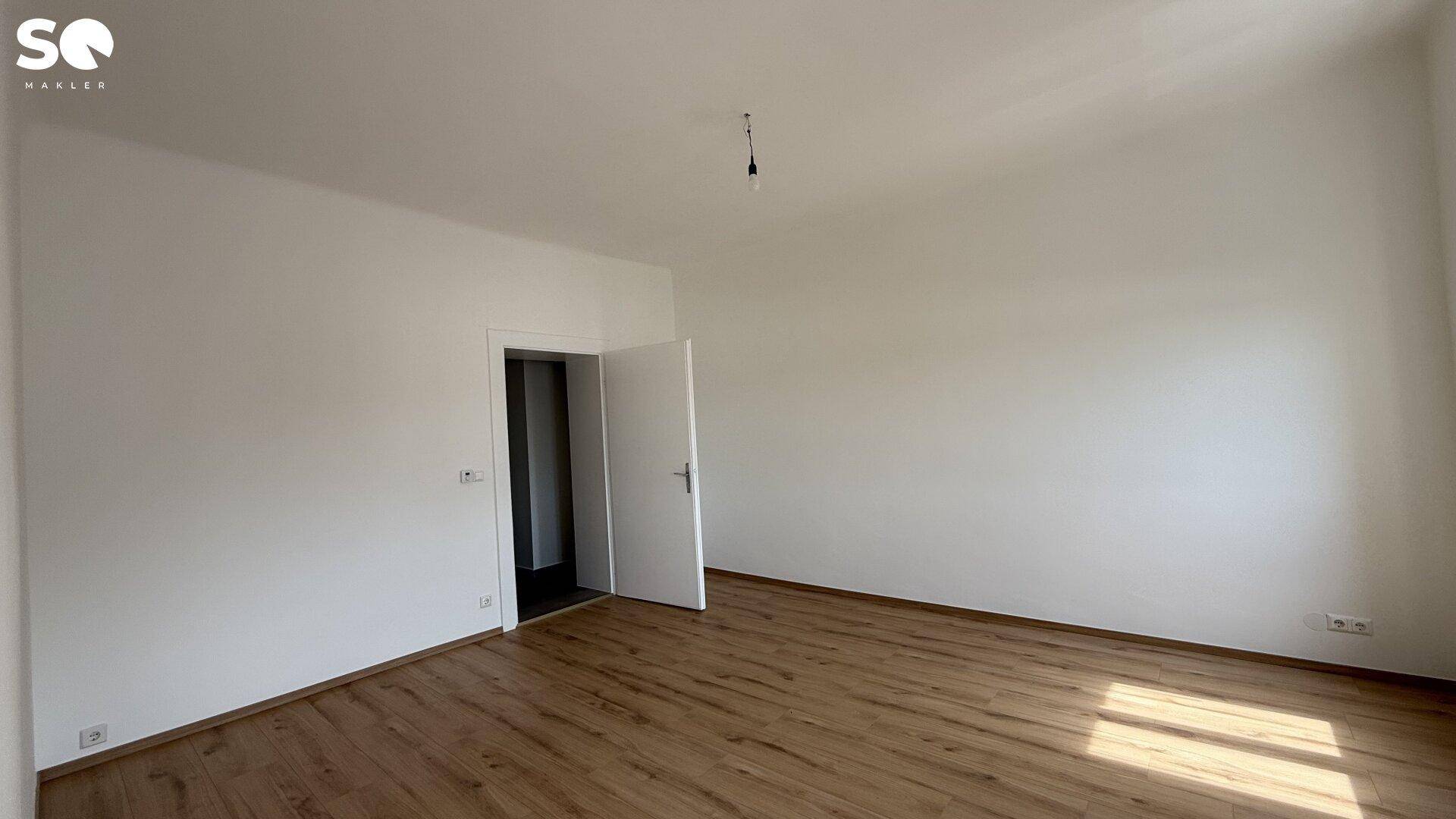 Leeres Zimmer mit Holzboden und offener Tür, die in einen angrenzenden Bereich führt.
