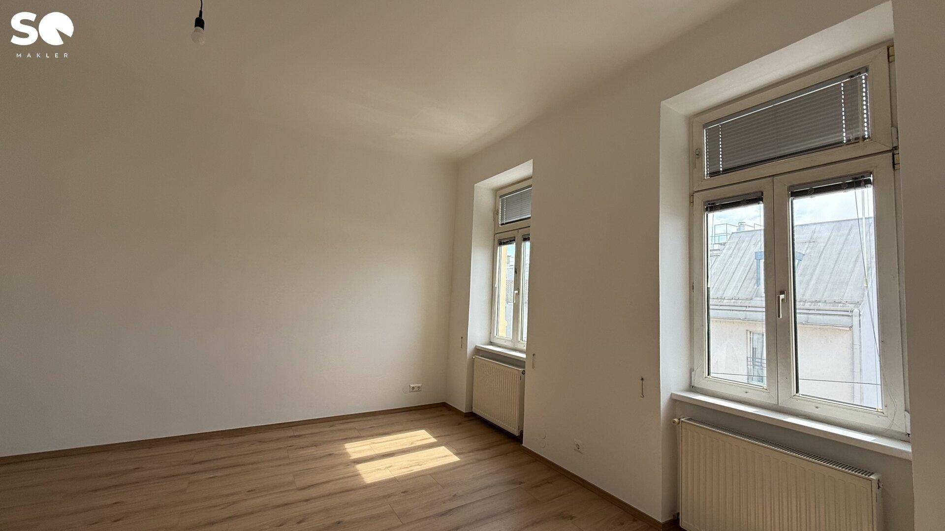 Helles, leeres Zimmer mit Holzfußboden und zwei Fenstern, bereit zur individuellen Gestaltung.