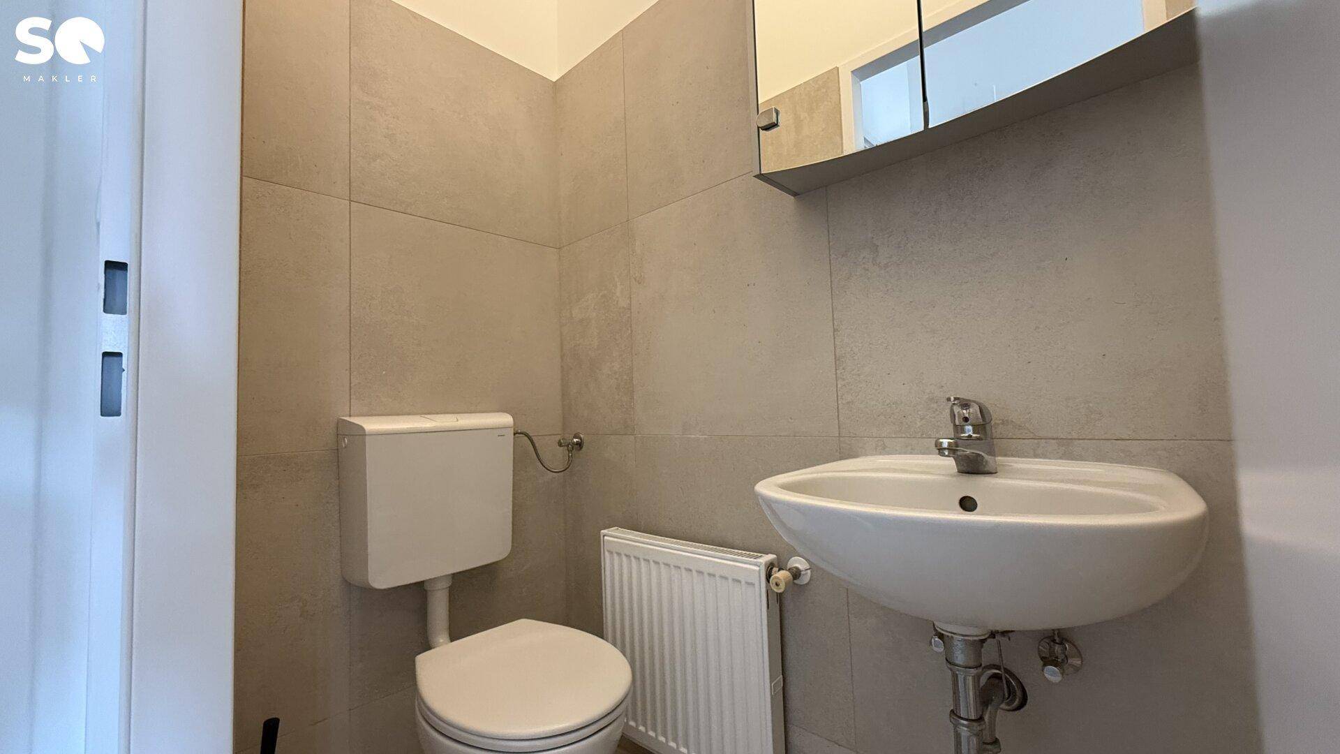 Sauberer Toilettenraum mit Waschbecken, WC und Heizung, in hellen Fliesen gehalten.