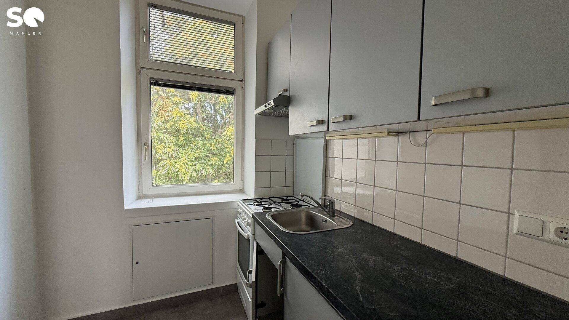 Kleine Küche mit grauen Schränken, weißem Fliesenspiegel und Fenster mit Blick ins Grüne.
