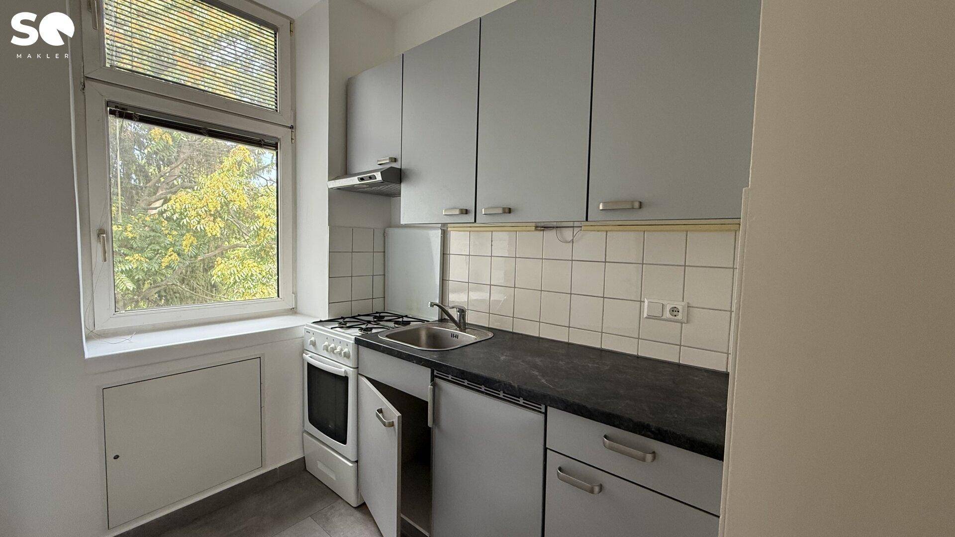 Einfache Küche mit grauen Schränken, Spüle und Gasherd, mit Fenster zum Garten.