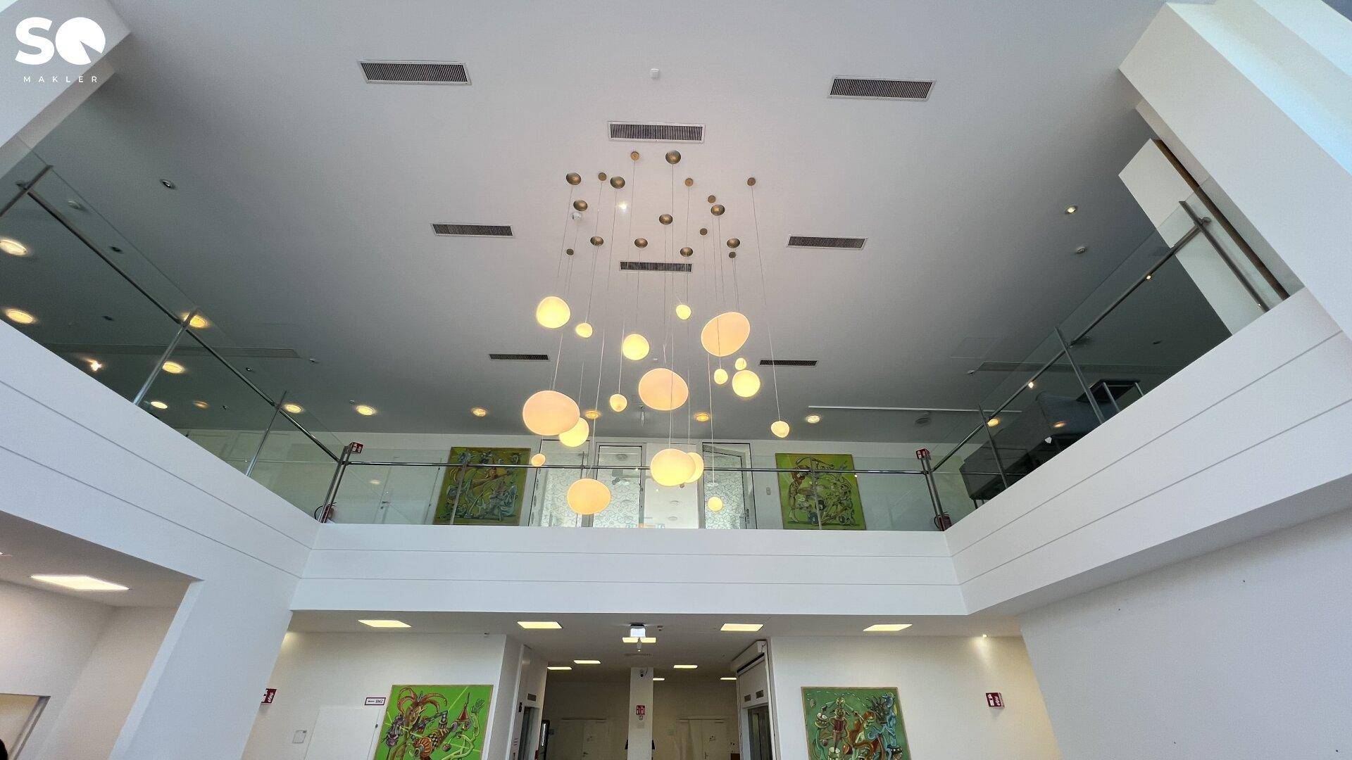Hohe Lobby mit beeindruckender Deckenleuchte, Glasgeländer und Kunstwerken im oberen Bereich.
