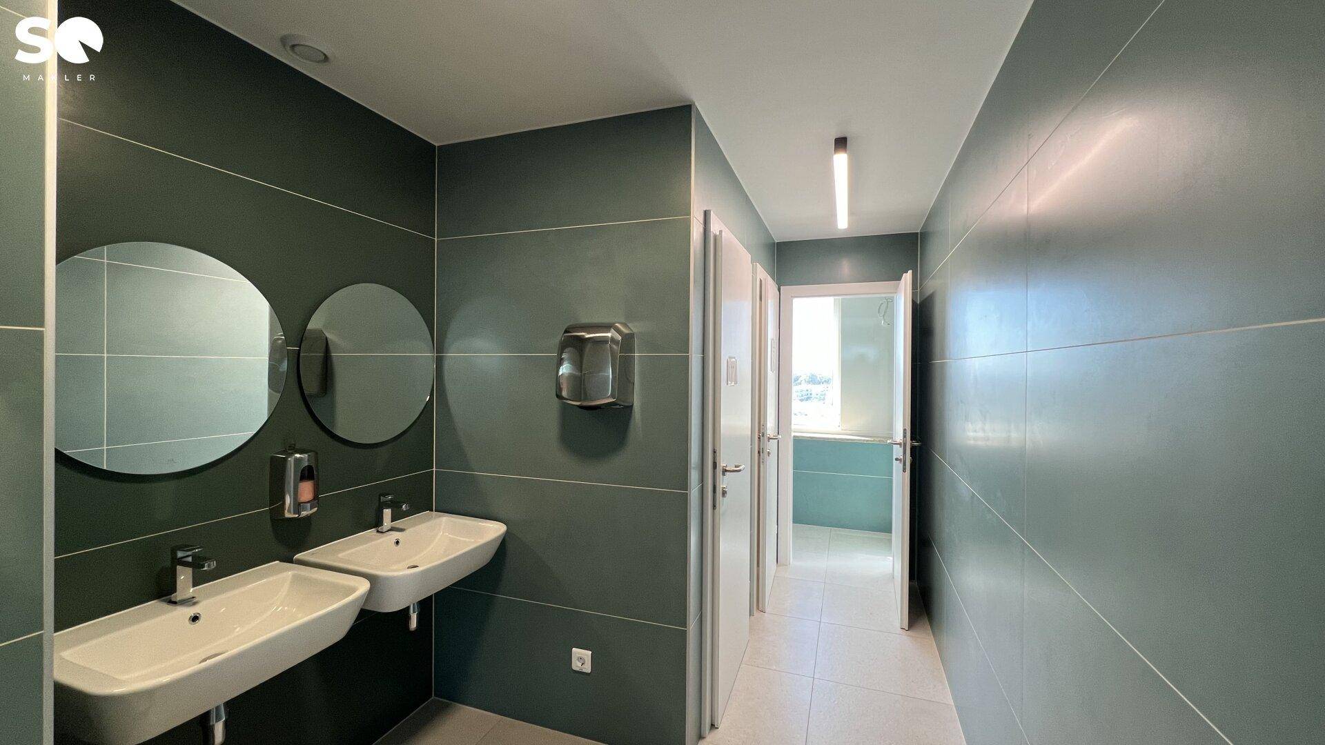 Moderner Waschraum mit zwei Waschbecken, runden Spiegeln und grünen Fliesen an den Wänden.