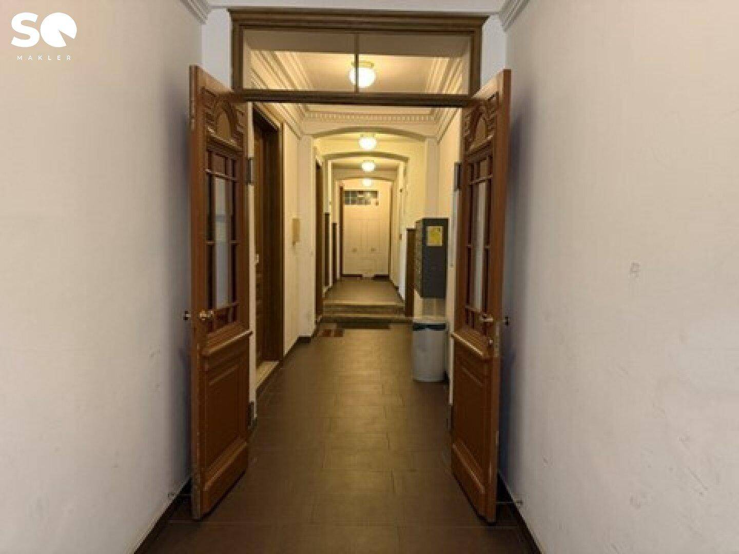 Eingangsbereich eines Altbaus mit hohen Decken, Stuck und Holztüren, der einen langen Flur mit weiteren Türen zeigt.